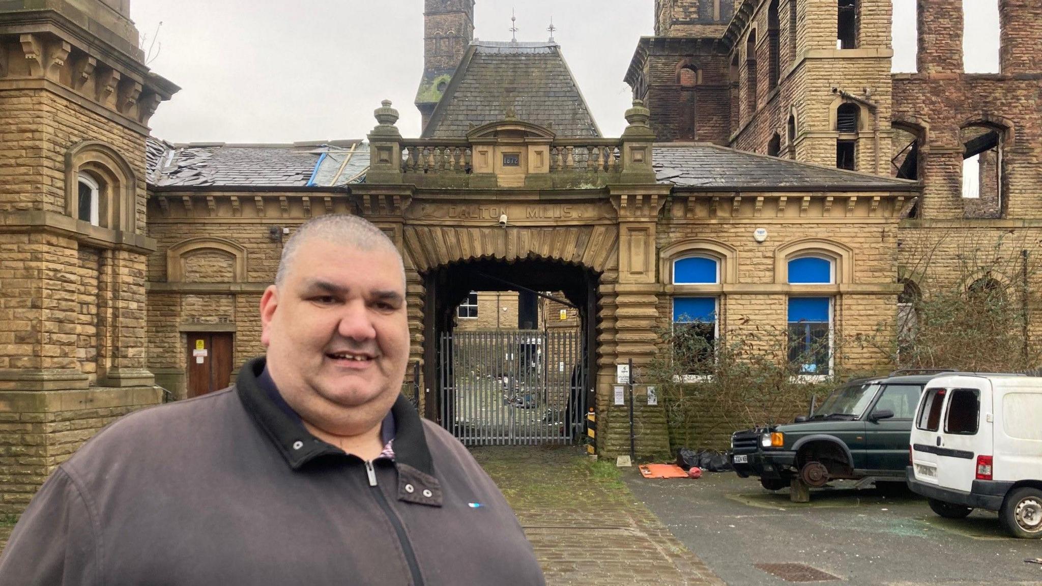 A shaven-headed man wearing a dark, faded purple jacket with a dirty yellow sandstone archway in the background and two van, one with its axle on a brick, to one side.
