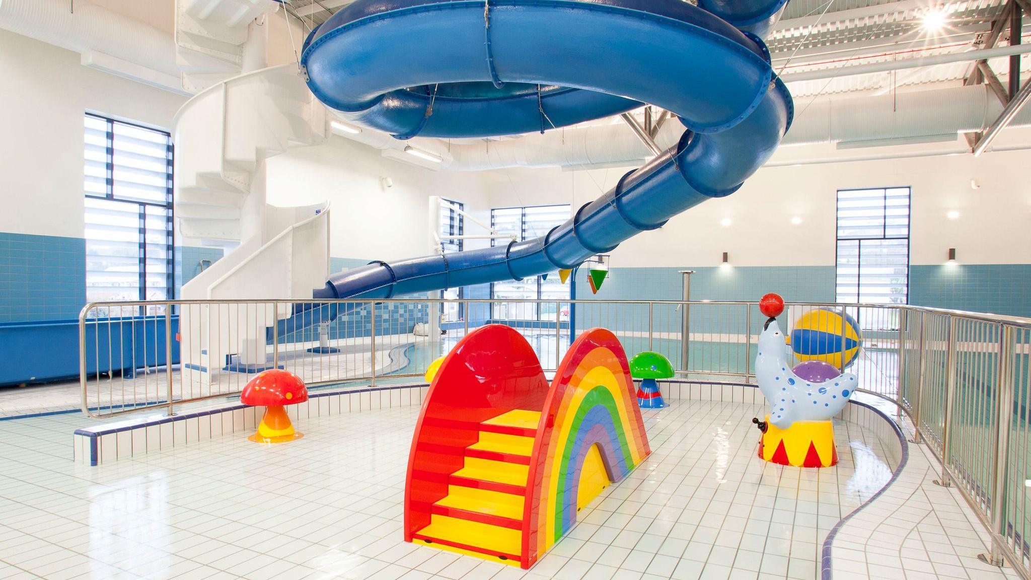 The children's play pool in Newry Leisure Centre.   It consists of a very shallow, white-tiled pool situated under a large blue flume which coils up towards the ceiling.  Children's play equipment is installed in the pool, including a small slide in the shape of a rainbow, a circus seal playing with a ball and colourful mushrooms.