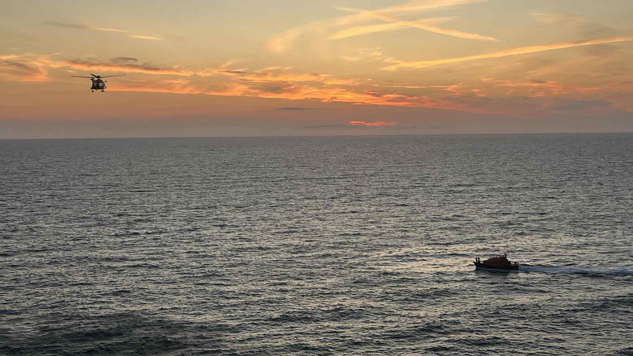 A helicopter and a lifeboat search the sea for a missing person during sunset.