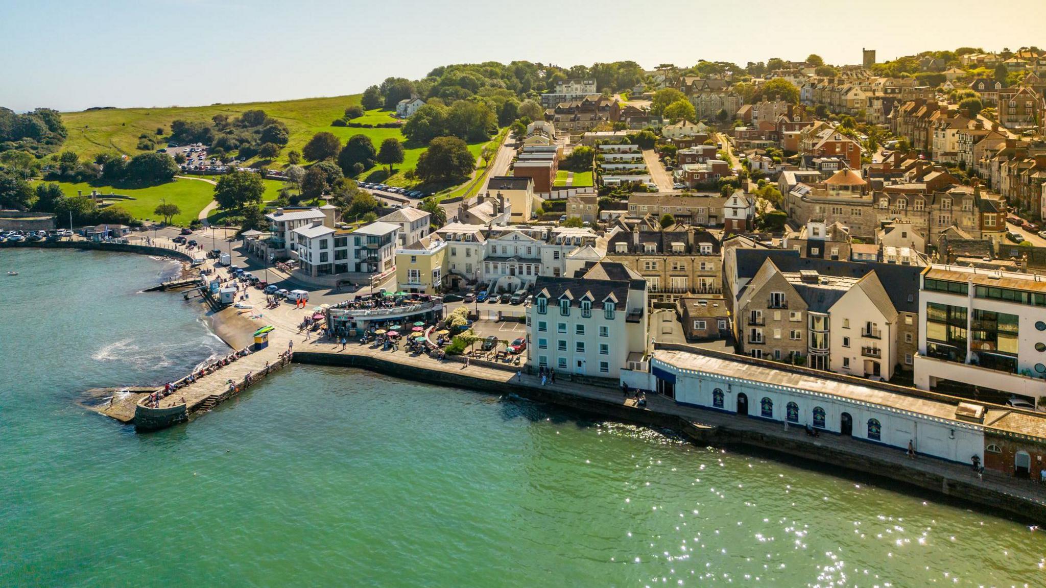 View of Swanage taken from an elevated position over the sea. There is a pier over some blue-green water in the foreground. Behind that are several residential buildings.