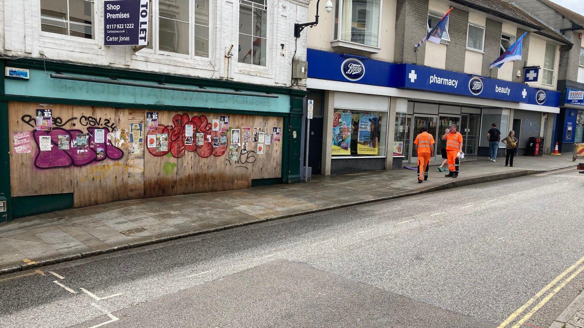 A wide shot of one of the shops on Market Jew Street which has been boarded up and covered with graffiti. There is a cleaning crew outside of it.