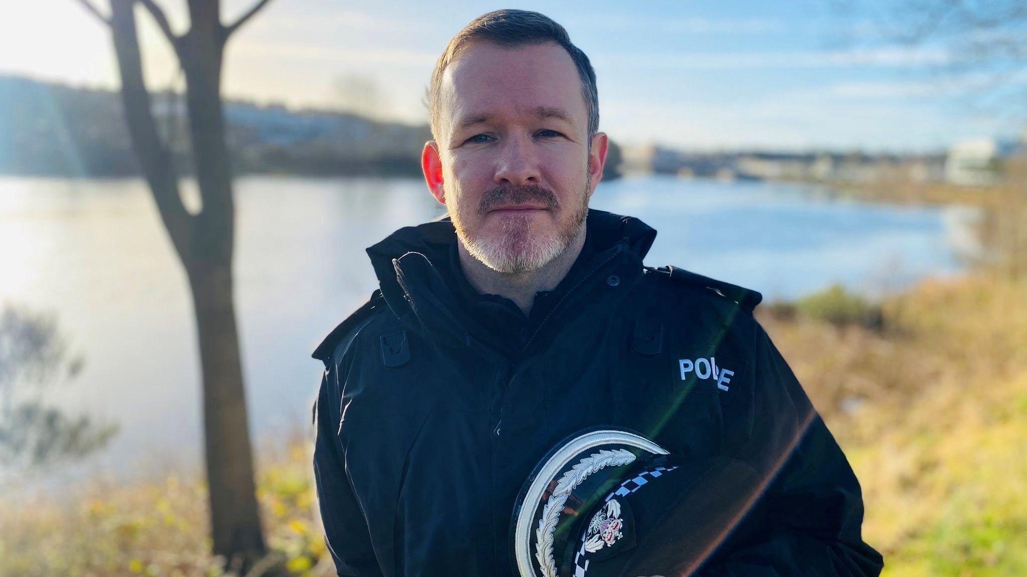 Police officer Supt Davie Howieson in black uniform and looking at camera, with a river and riverbank behind him