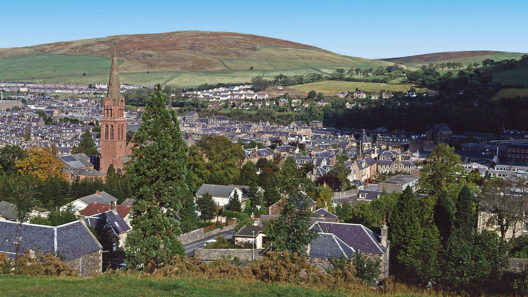 A general view of a town surrounded by green hills. There is a church steeple in the left of the picture.