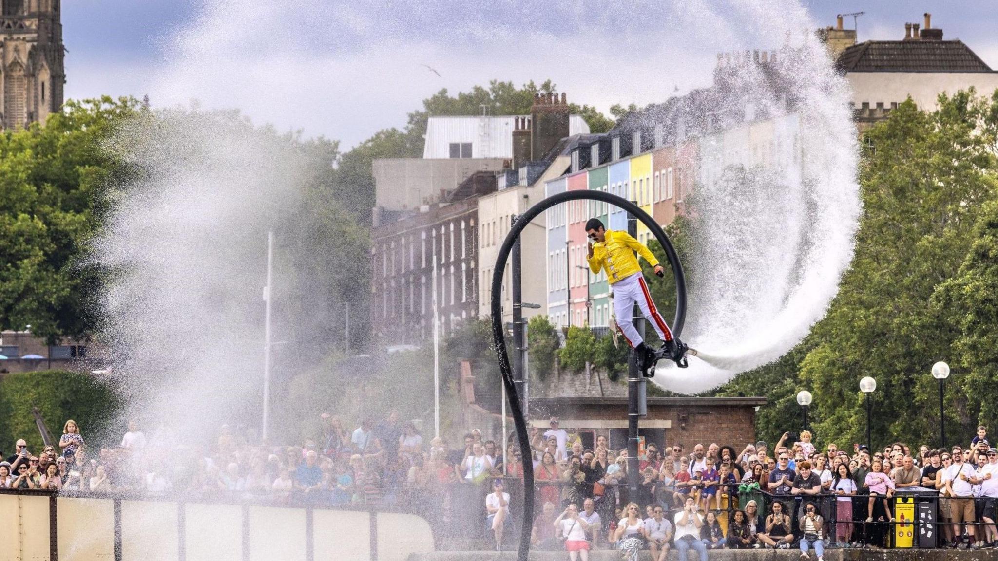 A man in a yellow outfit flies through the air using water-powered jets coming from hoses attached to his feet at the Bristol Harbour Festival.