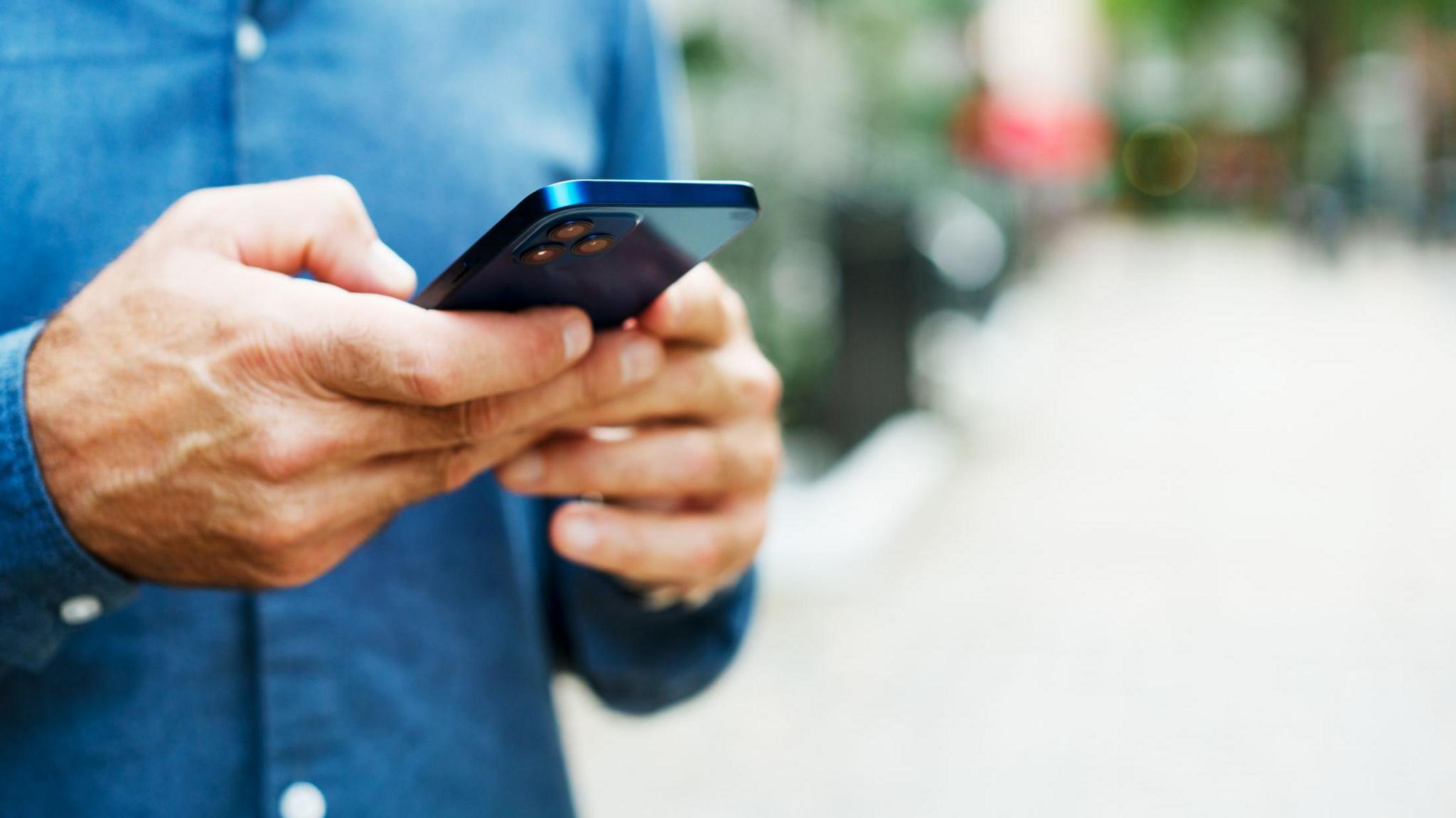 A man in a denim blue shirt holding a blue iPhone in both hands