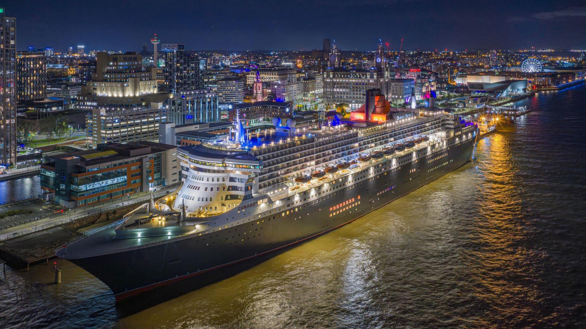 The Queen Mary 2 ocean liner as it arrives in Liverpool in front of the Liver Building. Liverpool city centre is lit up in the background.
