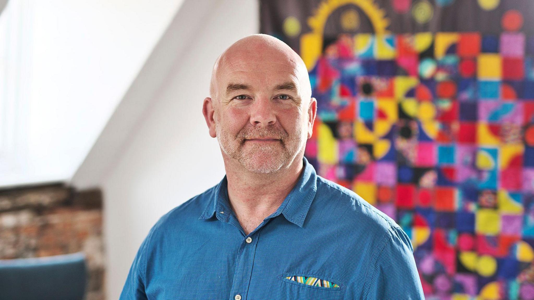 Clive Evans with very short white hair and beard, wearing a blue shirt and standing in front of a colourful abstract painting or fabric design.