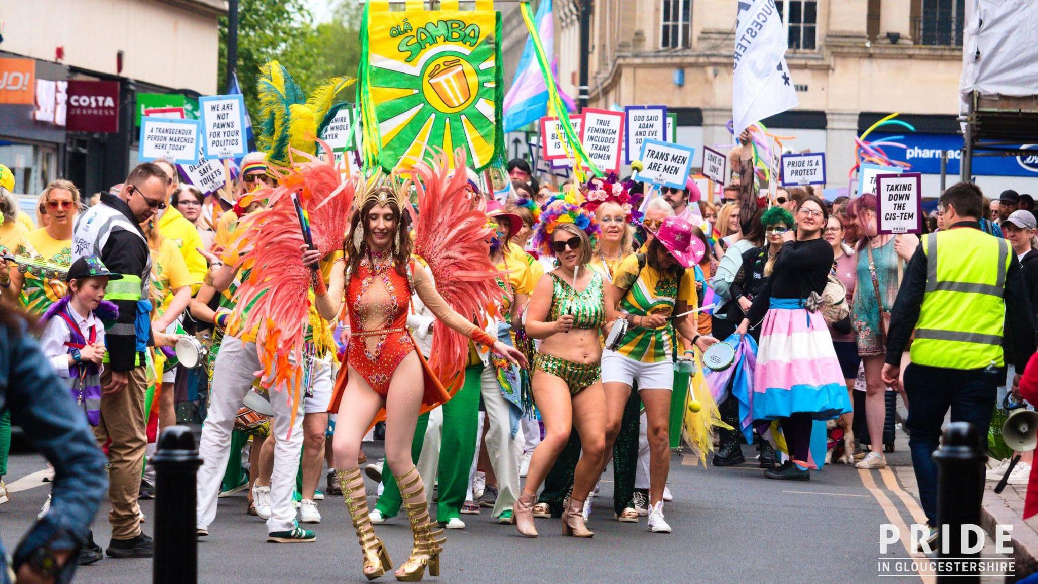 A Pride event featuring performers wearing multi-coloured outfits and carrying signs and banners in a town centre.