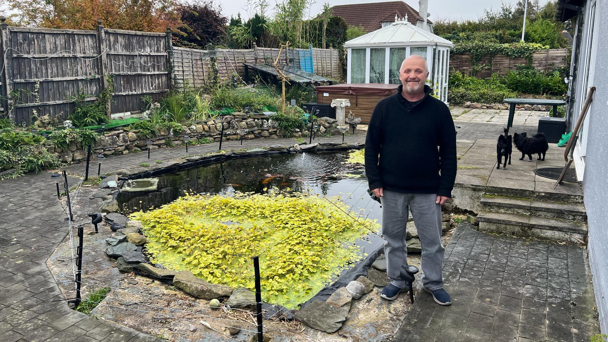 Nige Cooper standing beside his pond which has lots of green aquatic plants growing on top of it. The pond has an electric fence surrounding the water and his two chihuahuas are in the back view