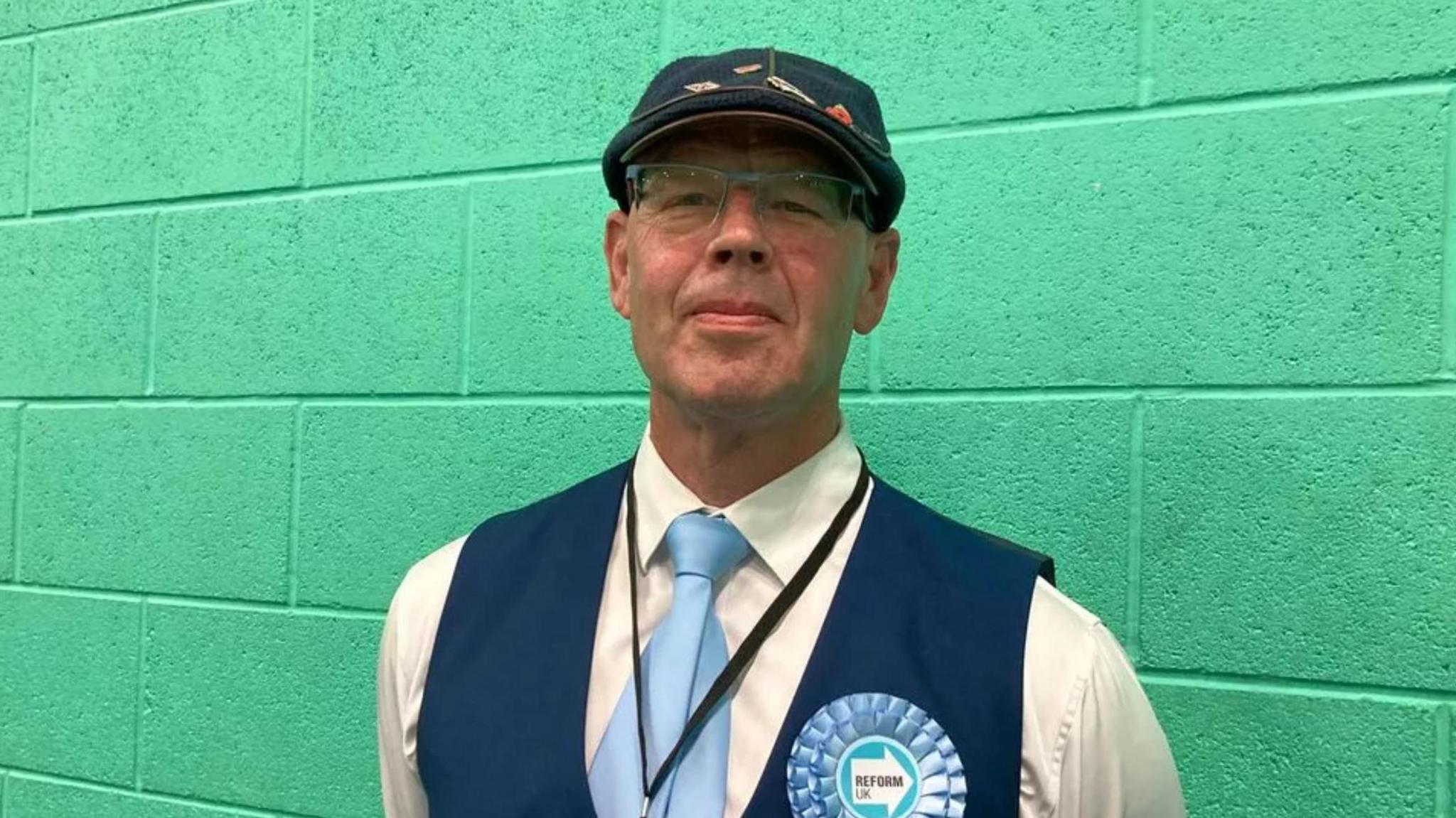 A man in a white shirt with blue waistcoat and blue cap and light blue tie and light blue Reform UK rosette standing against a green wall