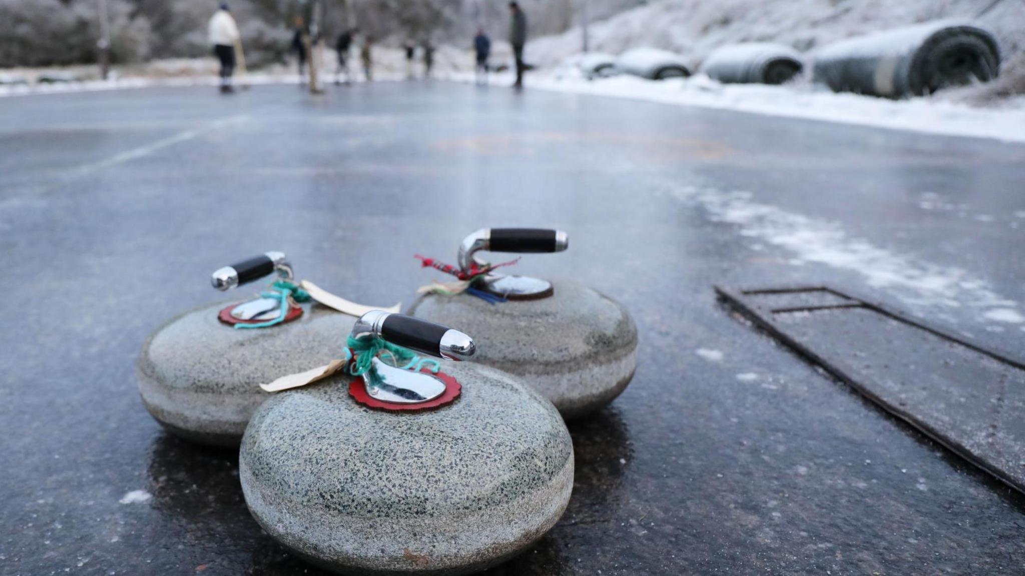 Three curling stones grouped together on the ice rink. In the distance and just out of focus are a group of curlers. There is snow lying around the edges of the rink, and large rolls of protective coverings for the rink.