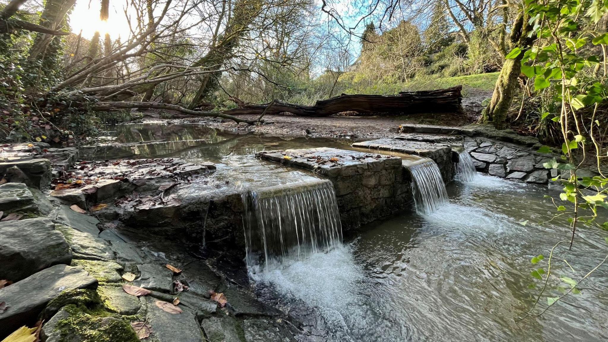 A set of stone steps pass over a small river in a park, with trees and grass behind in the background. The water is passing in small, clear waterfalls over three sets of stone gaps in the steps. It is a sunny day