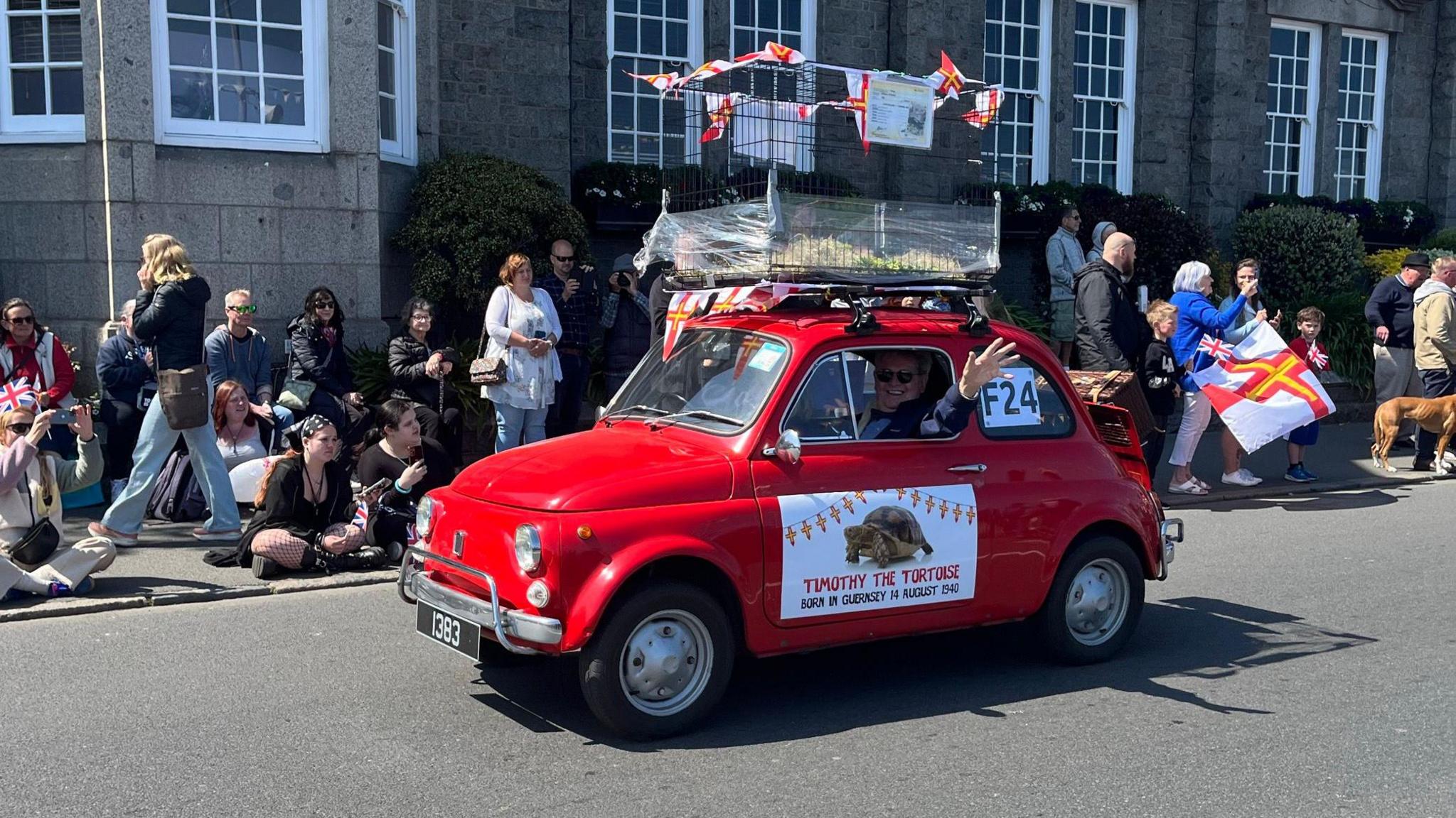 A picture of a red car with the tortoise in a cage on top of it. It has a logo on the side of the car with a picture of the animal. The crowds are lined up along the side of the streets.