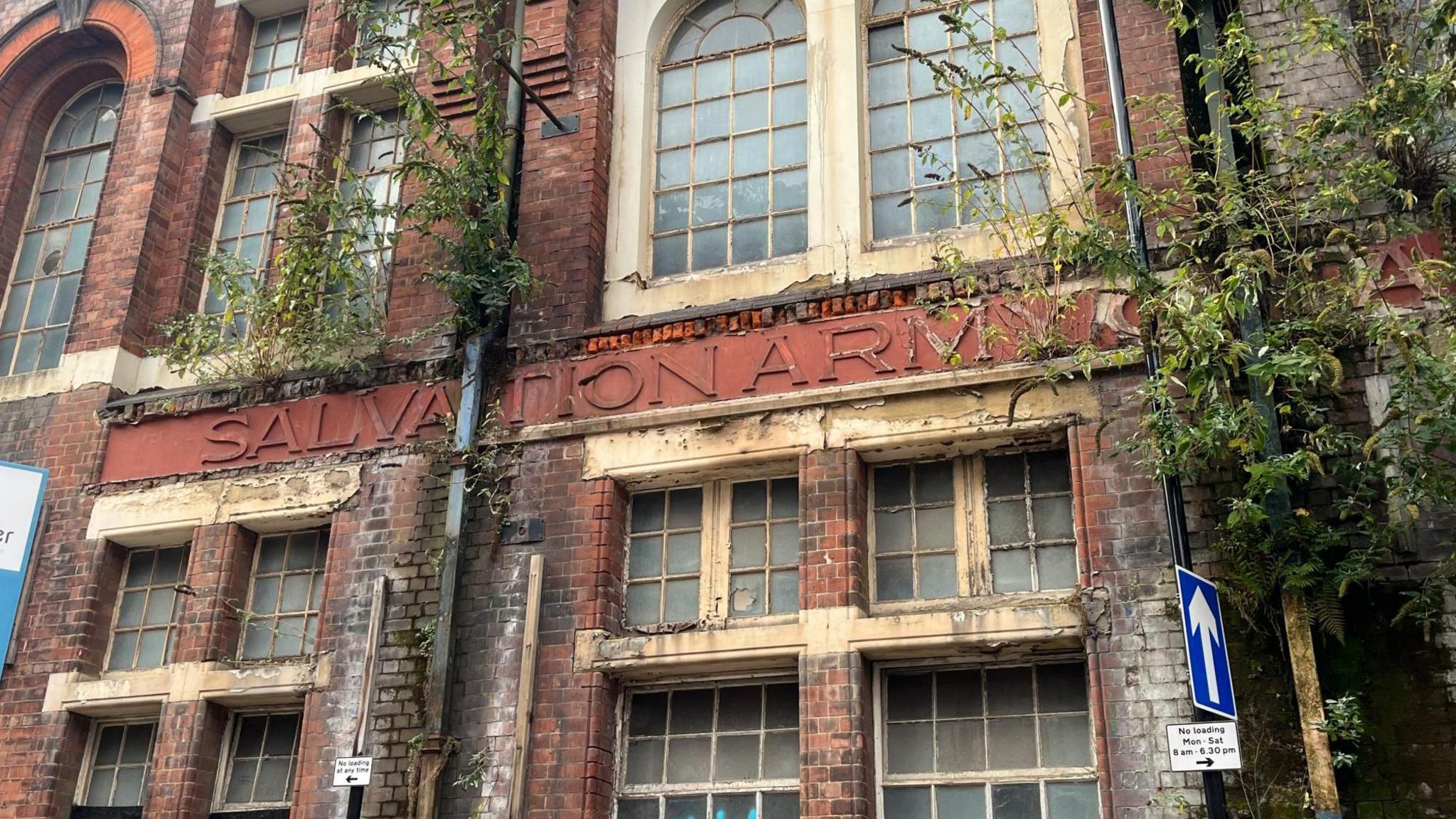 The red brick building with Salvation Army Citadel in the middle of the brickwork. There are trees and bushes growing out of the bricks making some of the working obscured