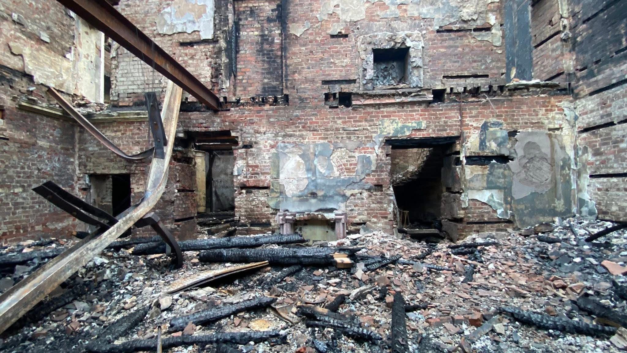 The image shows charred timbers and twisted metal littering the ground level of Woolton Hall after the fire. The ground is also littered with slates from the collapsed roof and everything that once covered the walls has been burned away to expose the brick