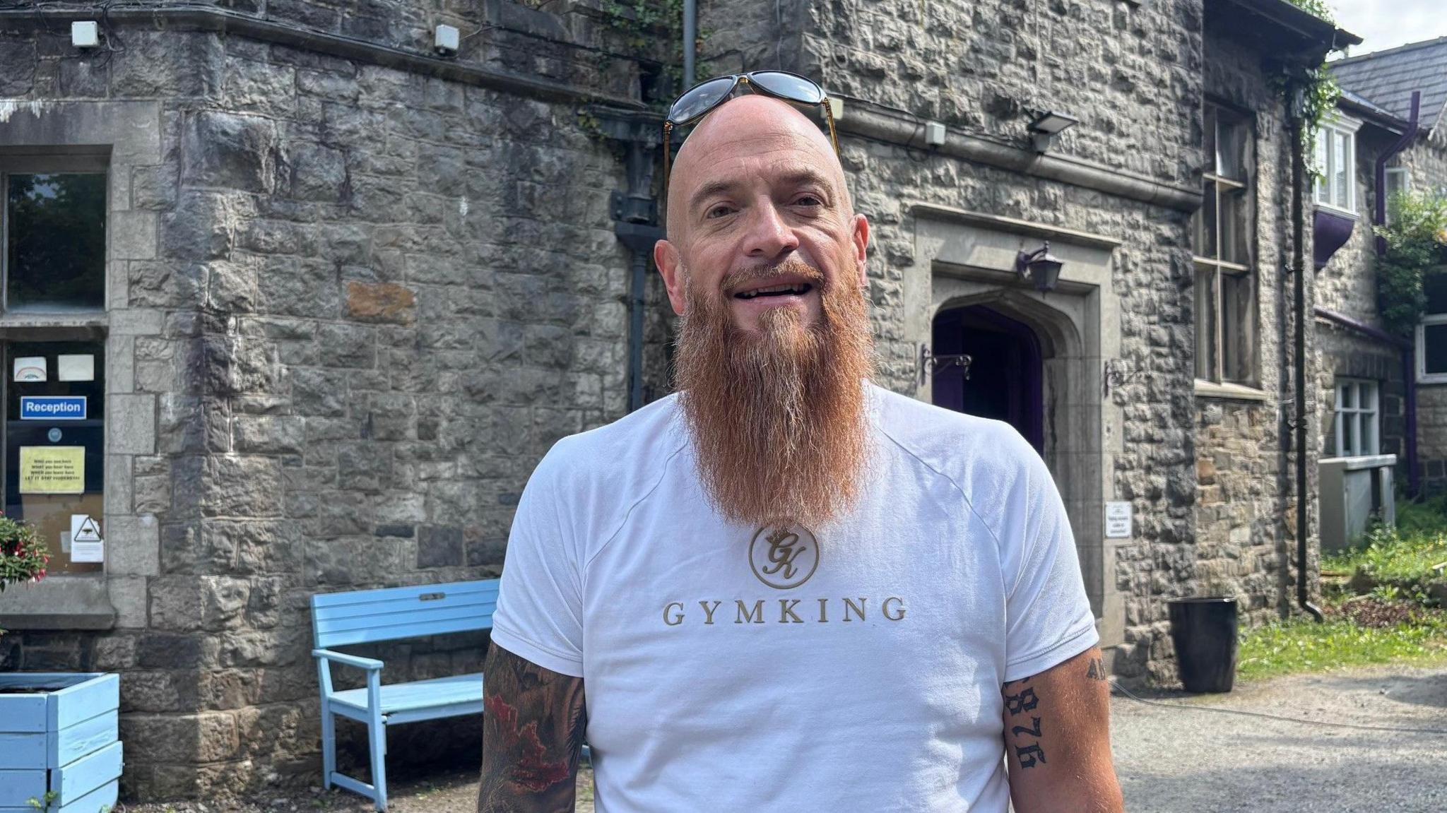 James Deakin stands outside historic large grey brick building with a light blue bench in front of it. He is bald with a long beard with sunglasses on his head and has a sleeve of tattoos.