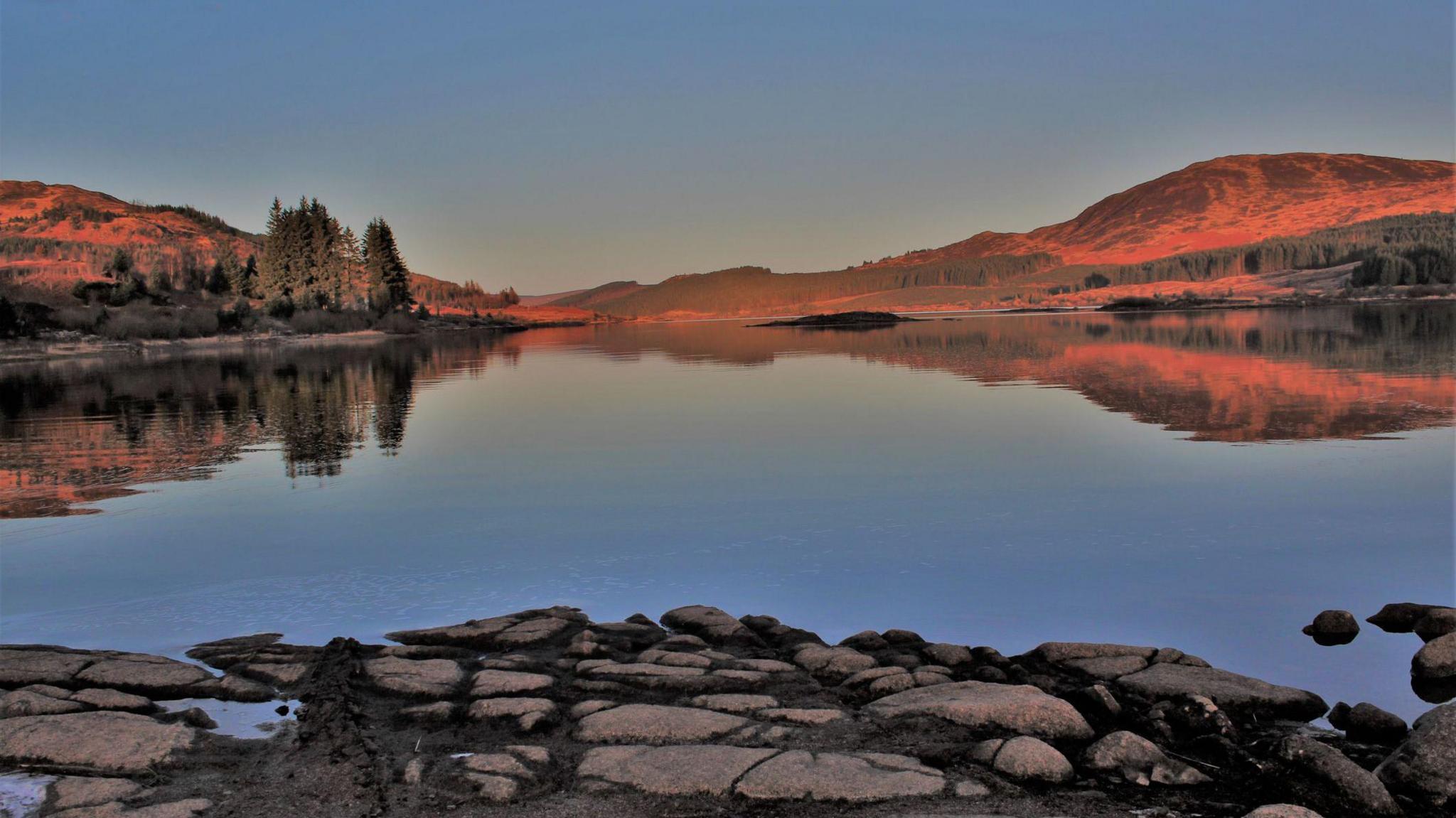 A scene across a Scottish loch with a stoney foreground and hills and trees in the background