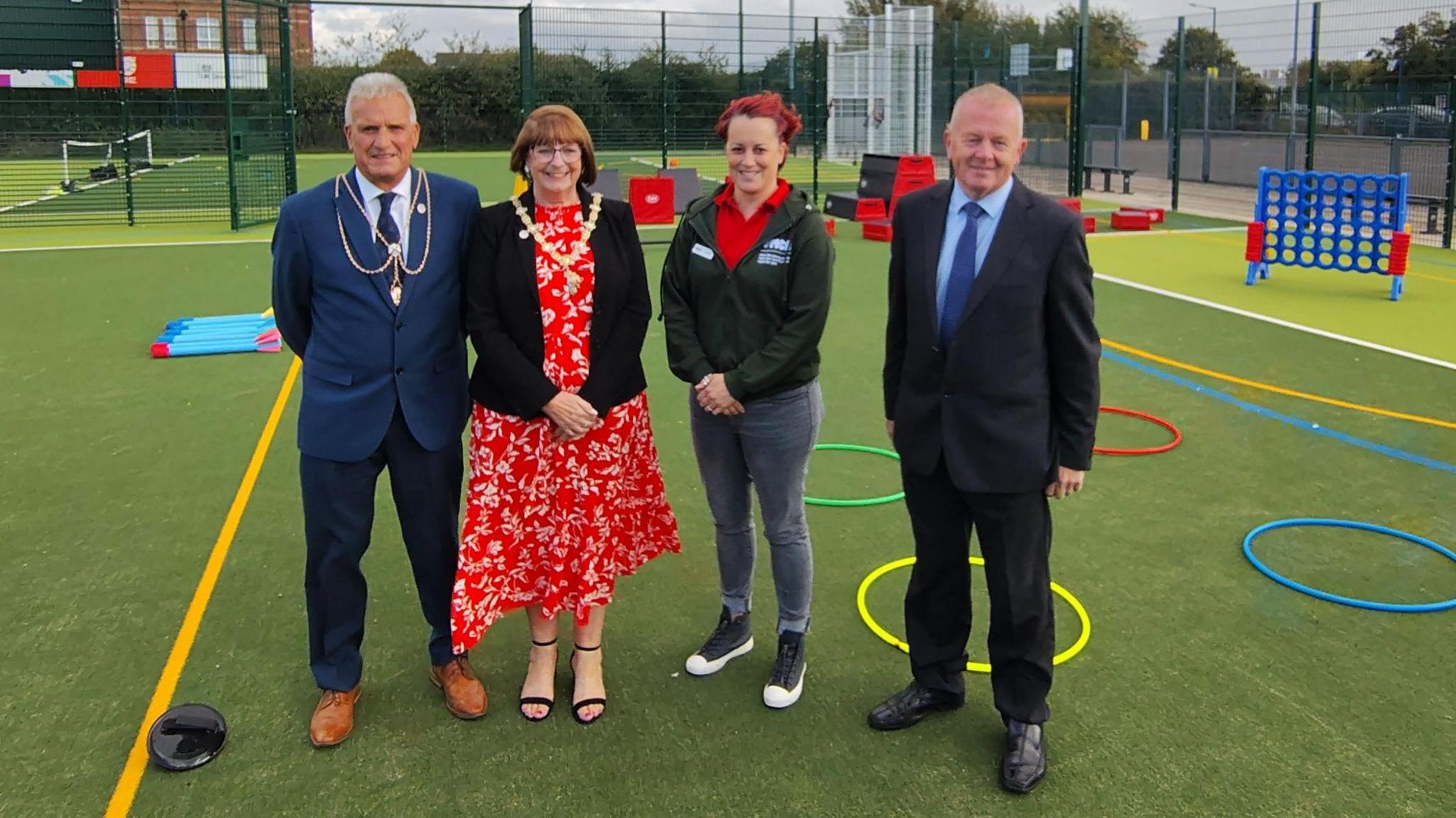 A number of people standing on one of the new sports pitches, There is a variety of equipment, including hoops and plastic barriers. The pitch has flood lights and is surrounded by a high mesh fence. 