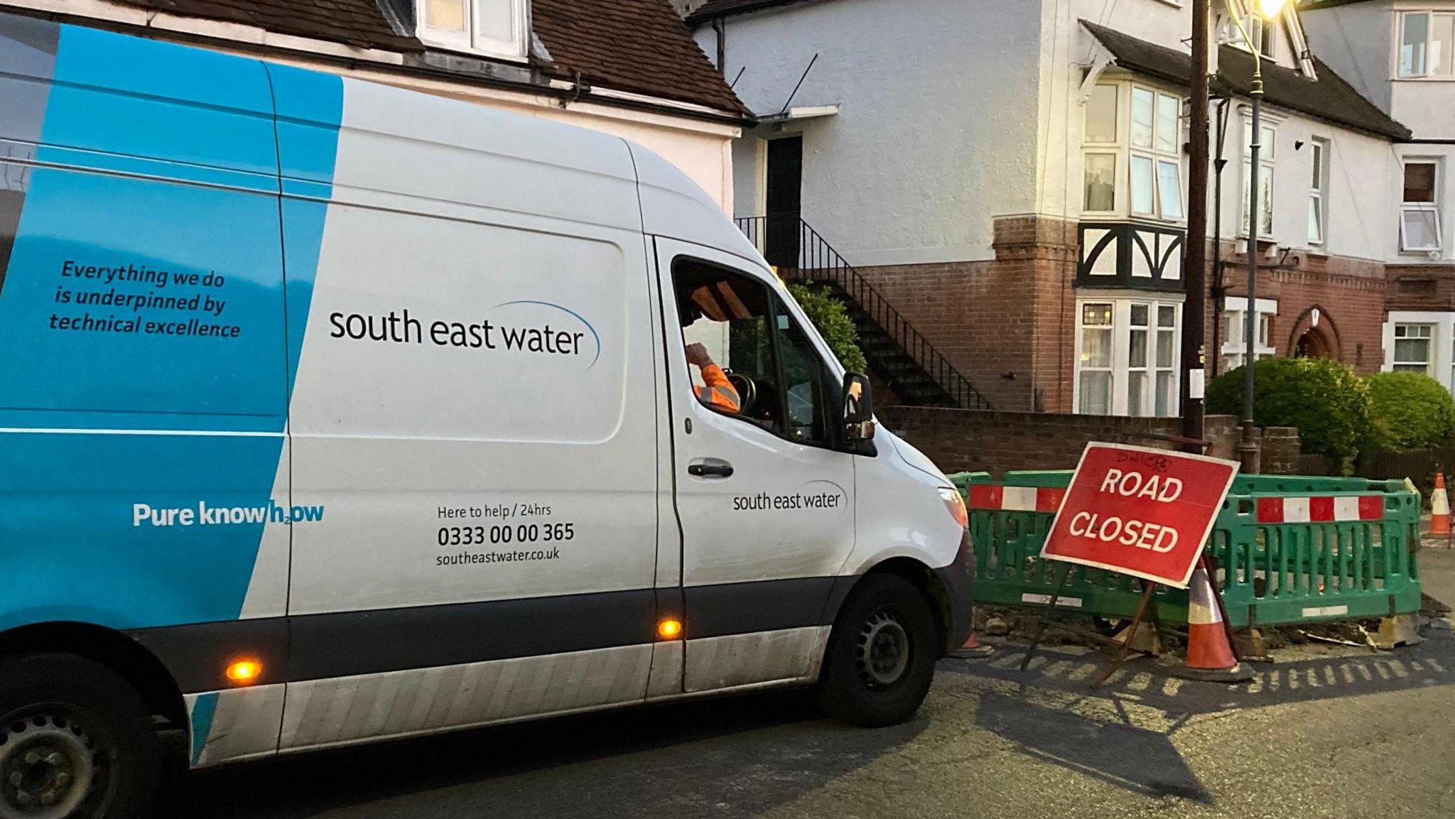 A blue and white SE Water van in front of a red sign reading "Road closed" with green fencing closing off an area of the street.