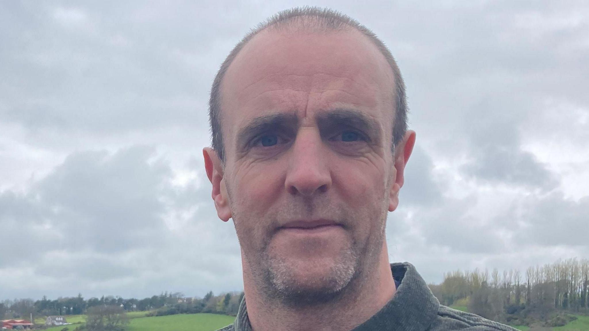 Mark H Durkan, in green shirt, with grass and trees in the background.
