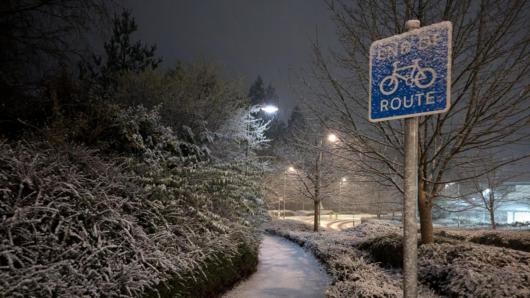 A snowy path with vegetation on either side with a light layer of snow. On the right, there's a blue sign signalling the end of the cycle route with snow on top.