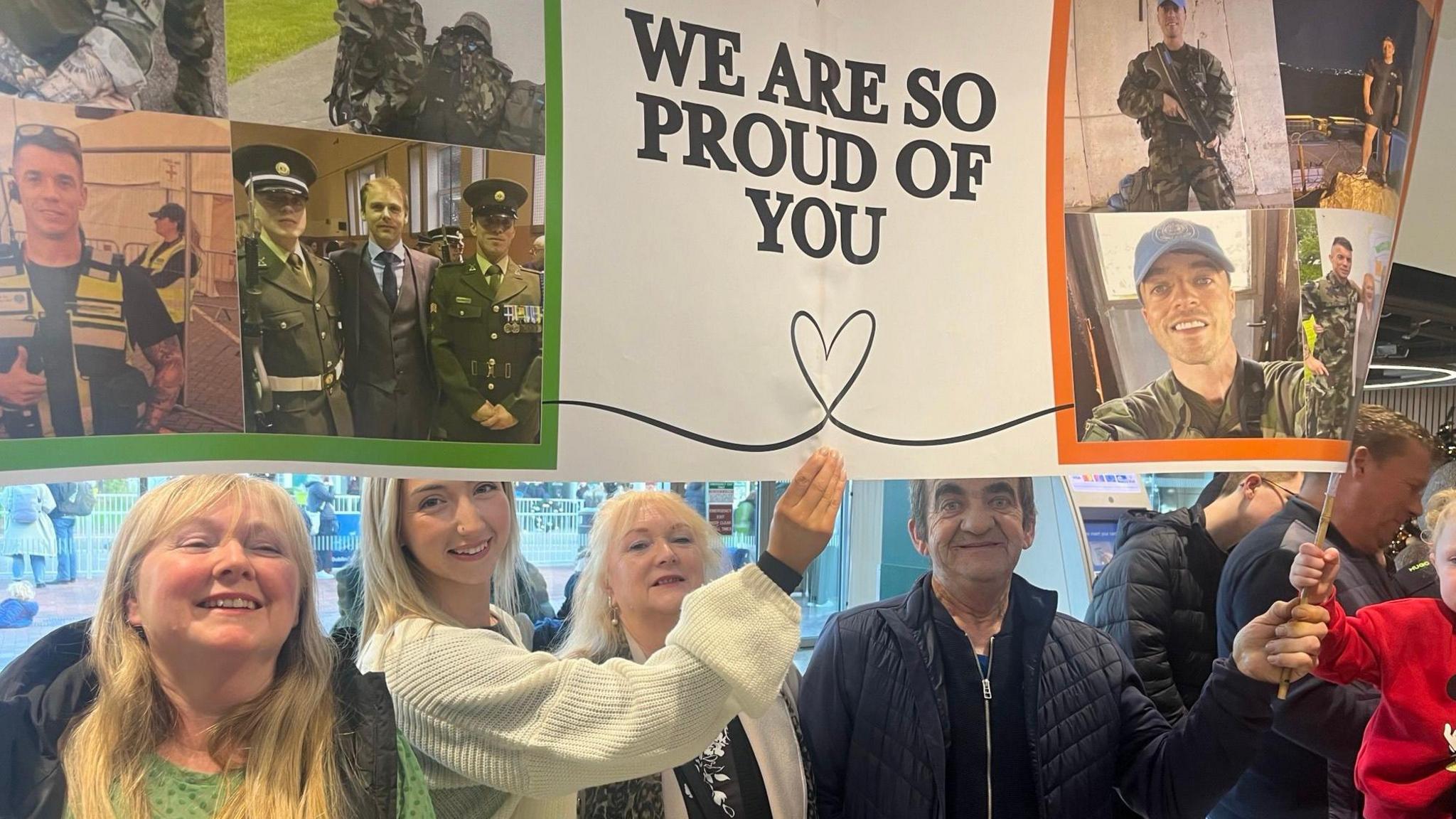 Three blonde women and one man hold up a banner that says - we are so proud of you. The banner also has a collage of two soldiers. 