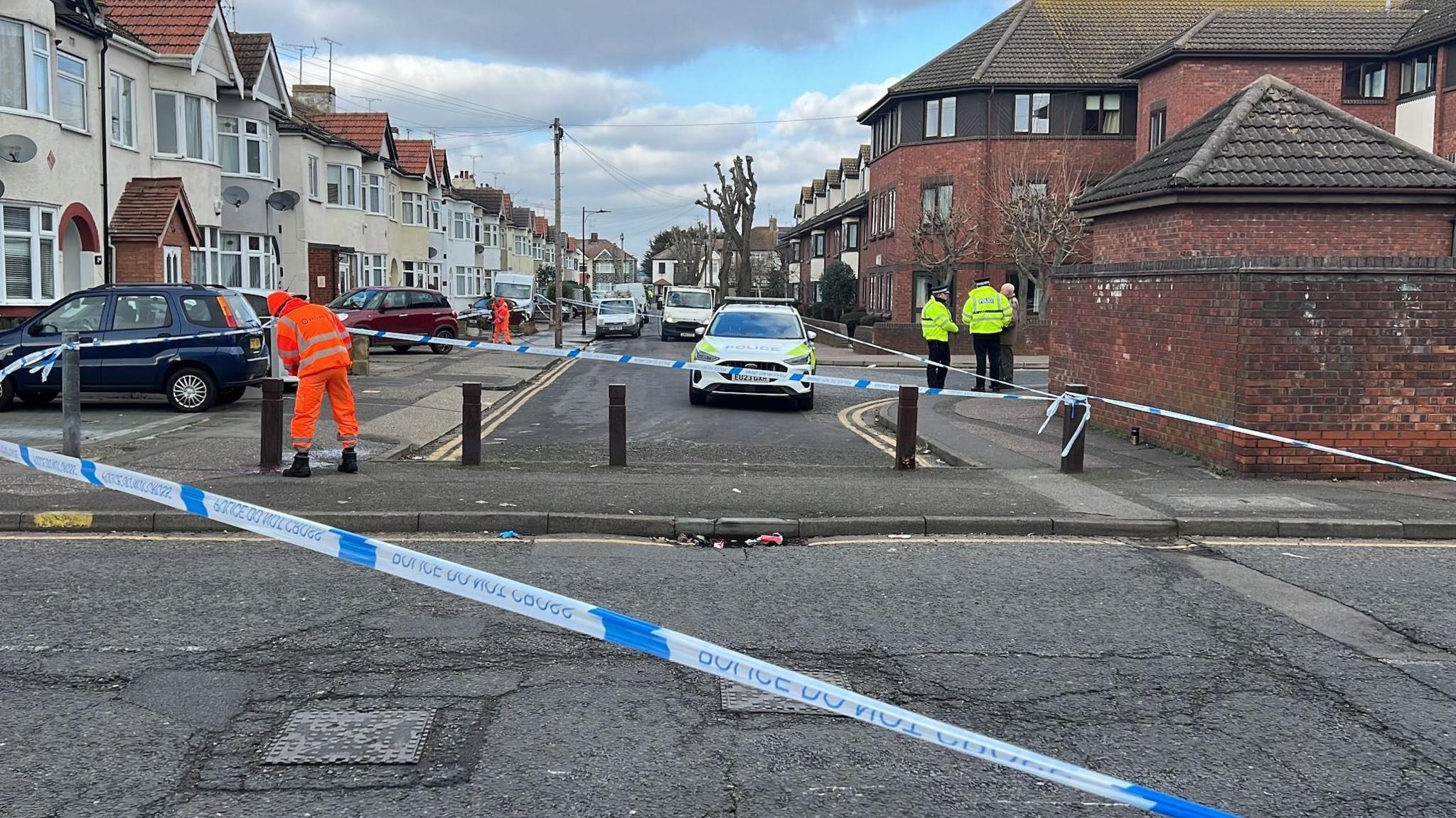 A police car at the end of a dead end road, with police tape across several roads. There are cars and a street with houses both side. Two police officers are taking to a man to the left and a man in orange high-vis is cleaning the street. 