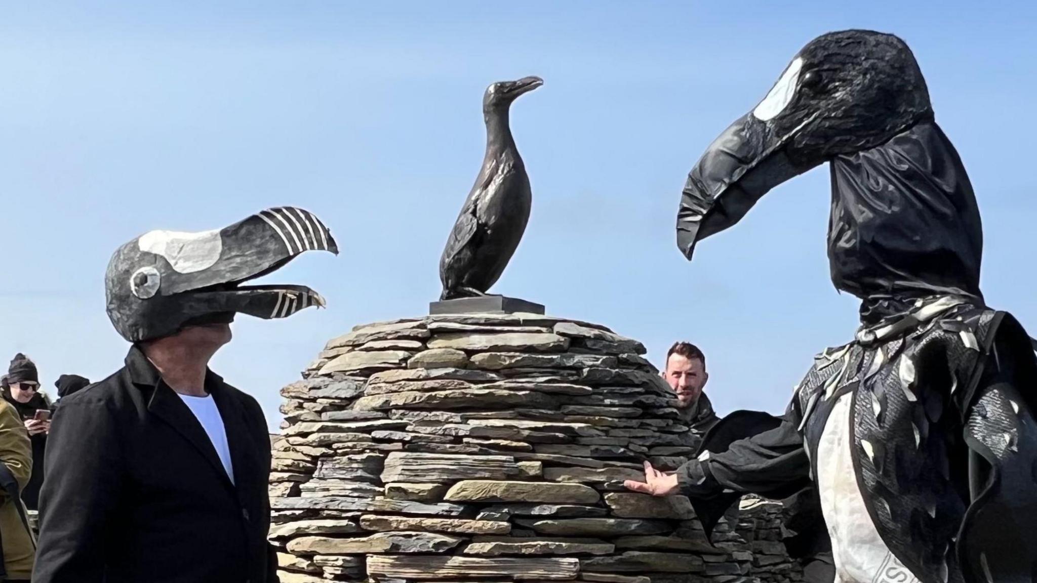 Two men dressed in black and white wearing headpieces modelled on the Great Auk stand next to an egg-shaped dry-stone cairn with a bronze statue of a Great Auk placed on top of it. Other people stand nearby.