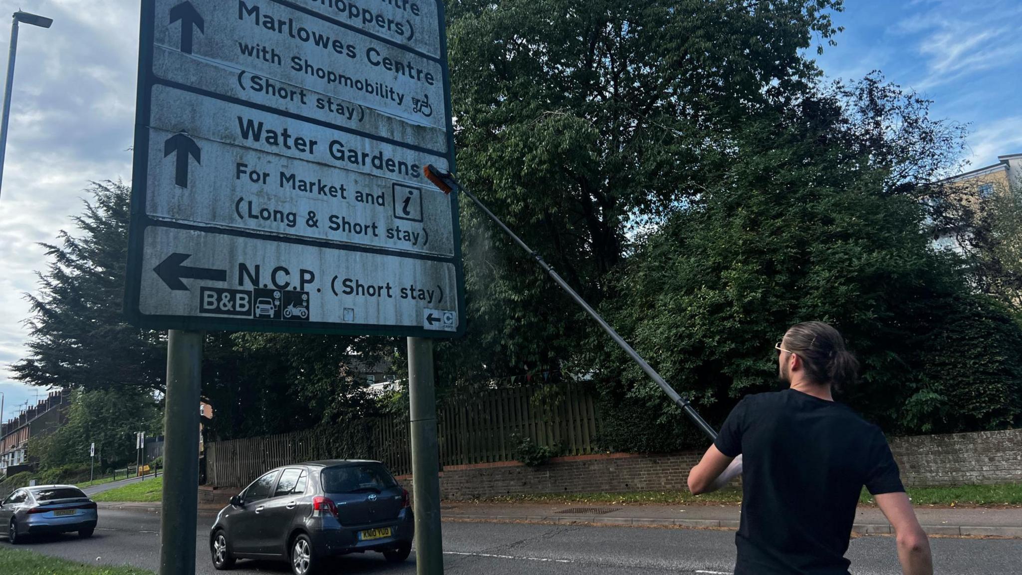 The Sign Guy, wearing a black t-shirt and sunglasses, is cleaning a large dirty road sign with directions using an extended pole brush.