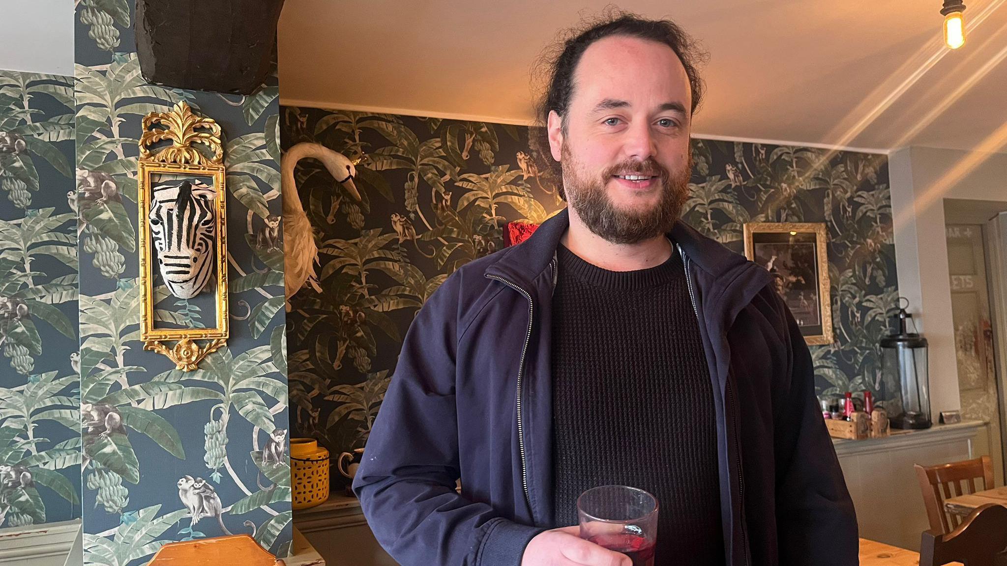 A bearded man stands in a pub holding a pint. He is wearing a black jumper and dark blue jacket. The pub is decorated with leafy wallpaper and has a porcelain zebra head hanging from the wall inside a gold photo frame. There is a similar porcelain swan also hanging from the wall. Wooden chairs and table can also be seen.