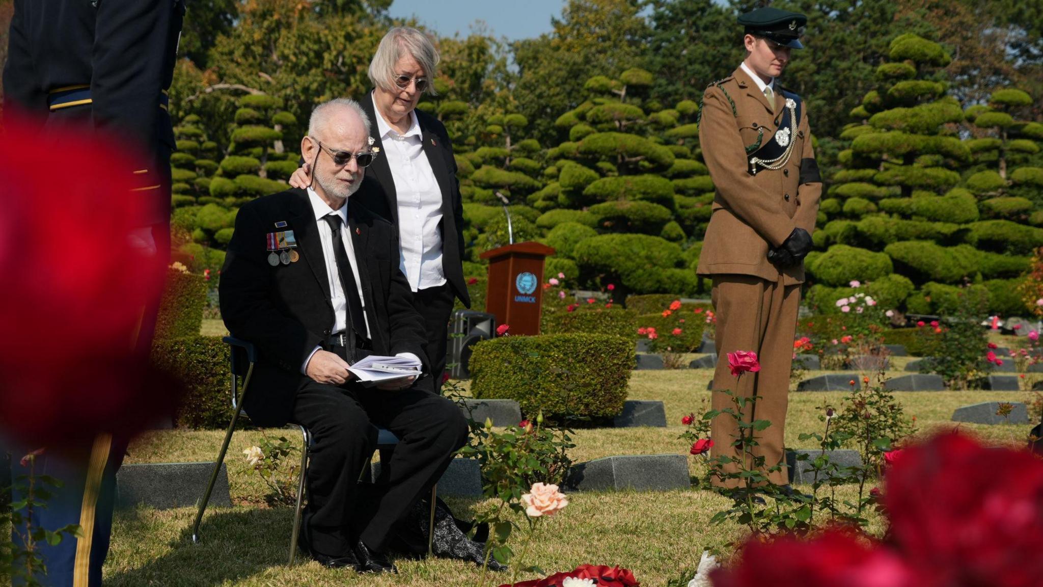 Michael Northey sits by his father's grave; a man in a wheelchair sits beside a grave in cemetery. A woman and a soldier stand behind him to his right.