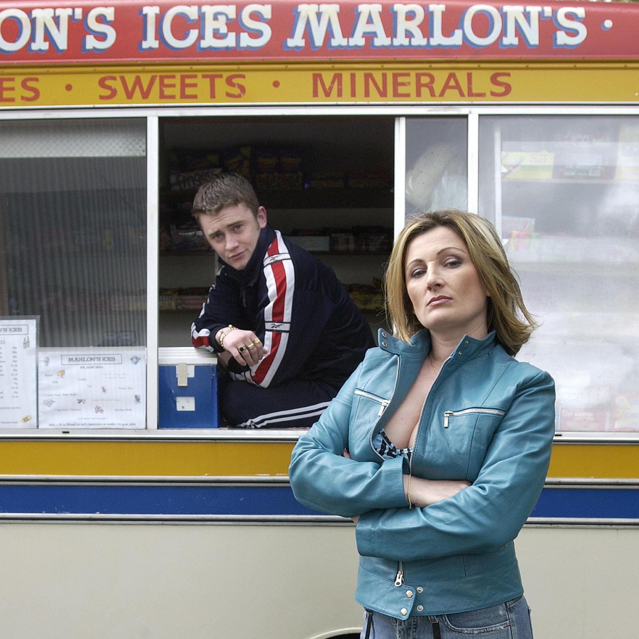 A person stands in the foreground with arms folded, wearing a turquoise jacket, patterned jeans, and holding a leopard‑print bag.
Behind them is a cream-and-yellow ice cream van with signage reading “Marlon’s Ices” and “Sweets • Minerals.”
Another person leans out of the serving window of the van, suggesting a casual street setting on a wet roadway.