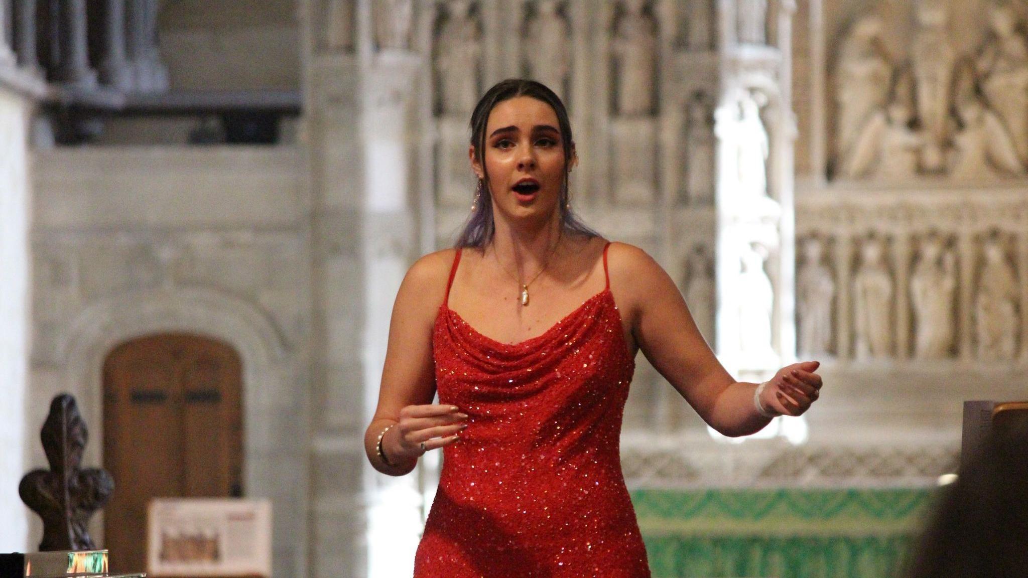 Megan Hastings singing opera in a church. Figures are carved into the stone behind her. She is wearing a red sparkly dress. She has long brown hair. 