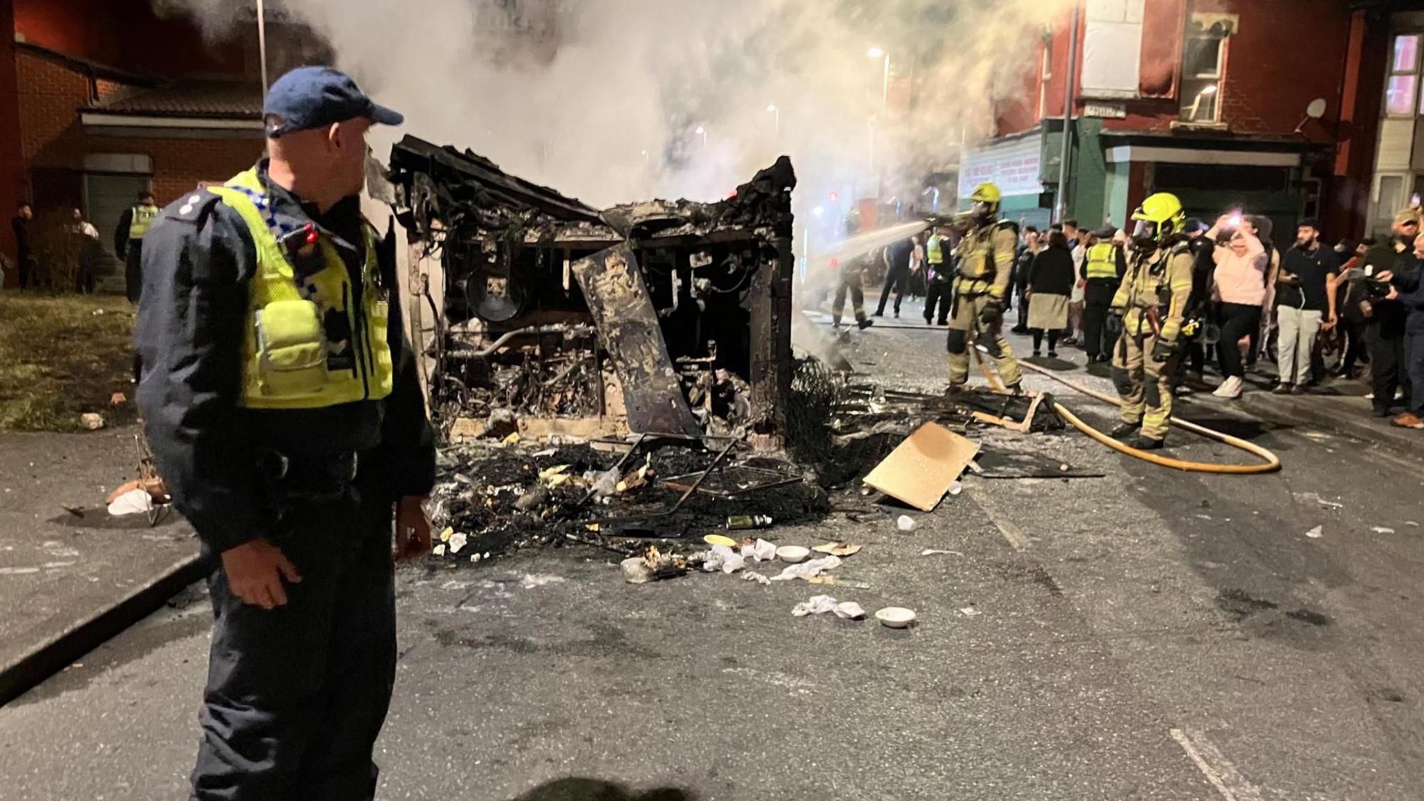 A police officer stands in front of a burnt-out bus