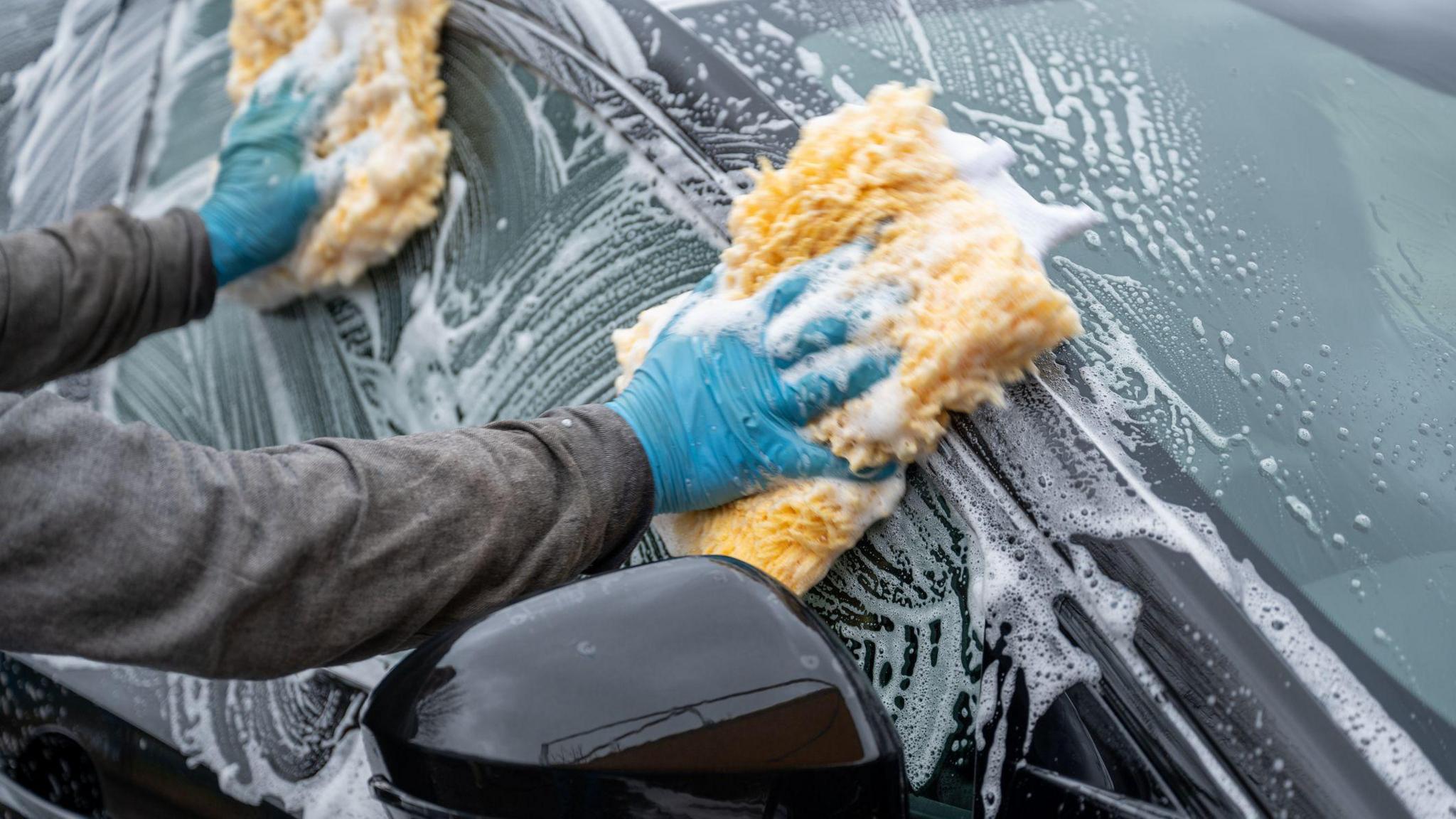 Two blue-gloved hands holding yellow sponges washing the window of a black car.