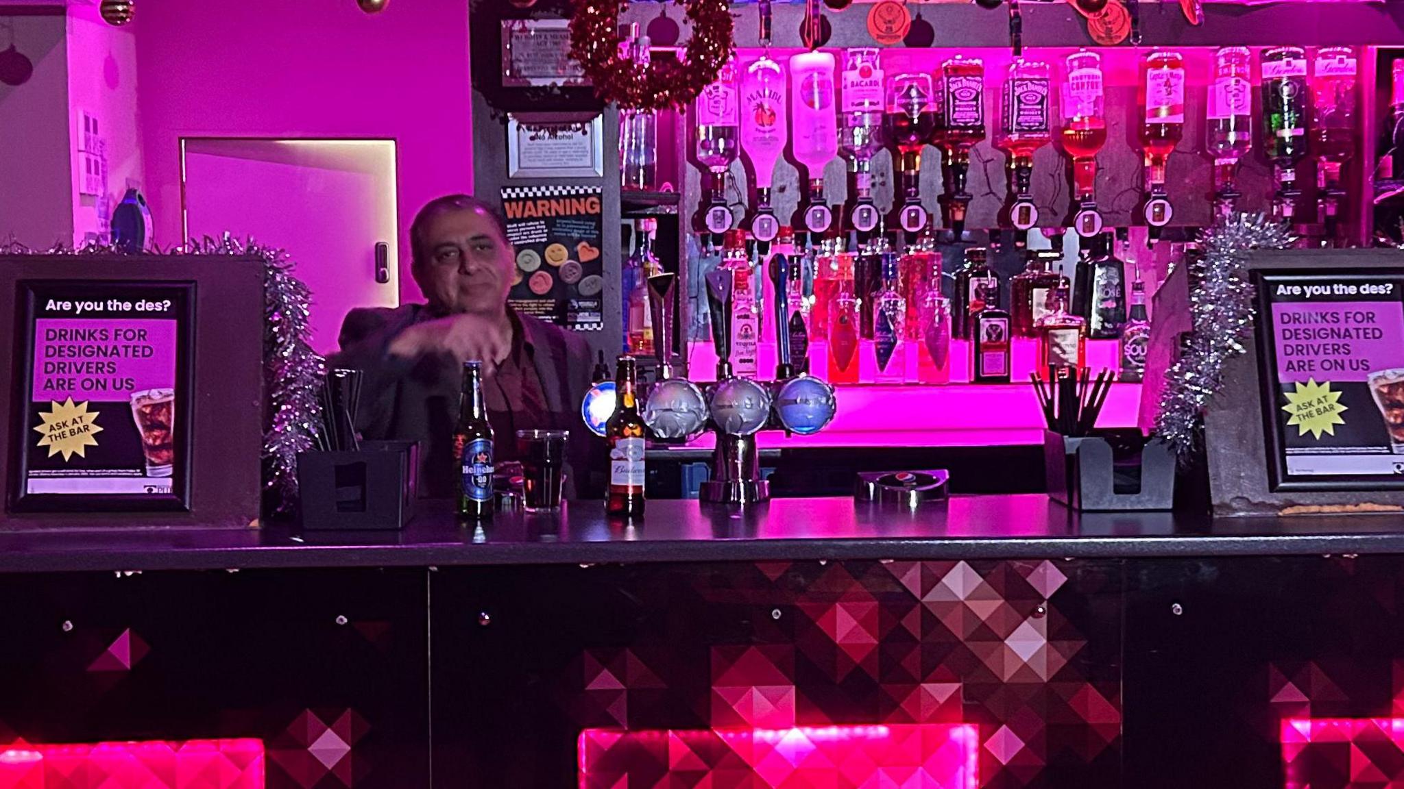 A bar area in purple ambient light with three red squares on the front. A man stands behind the bar. 