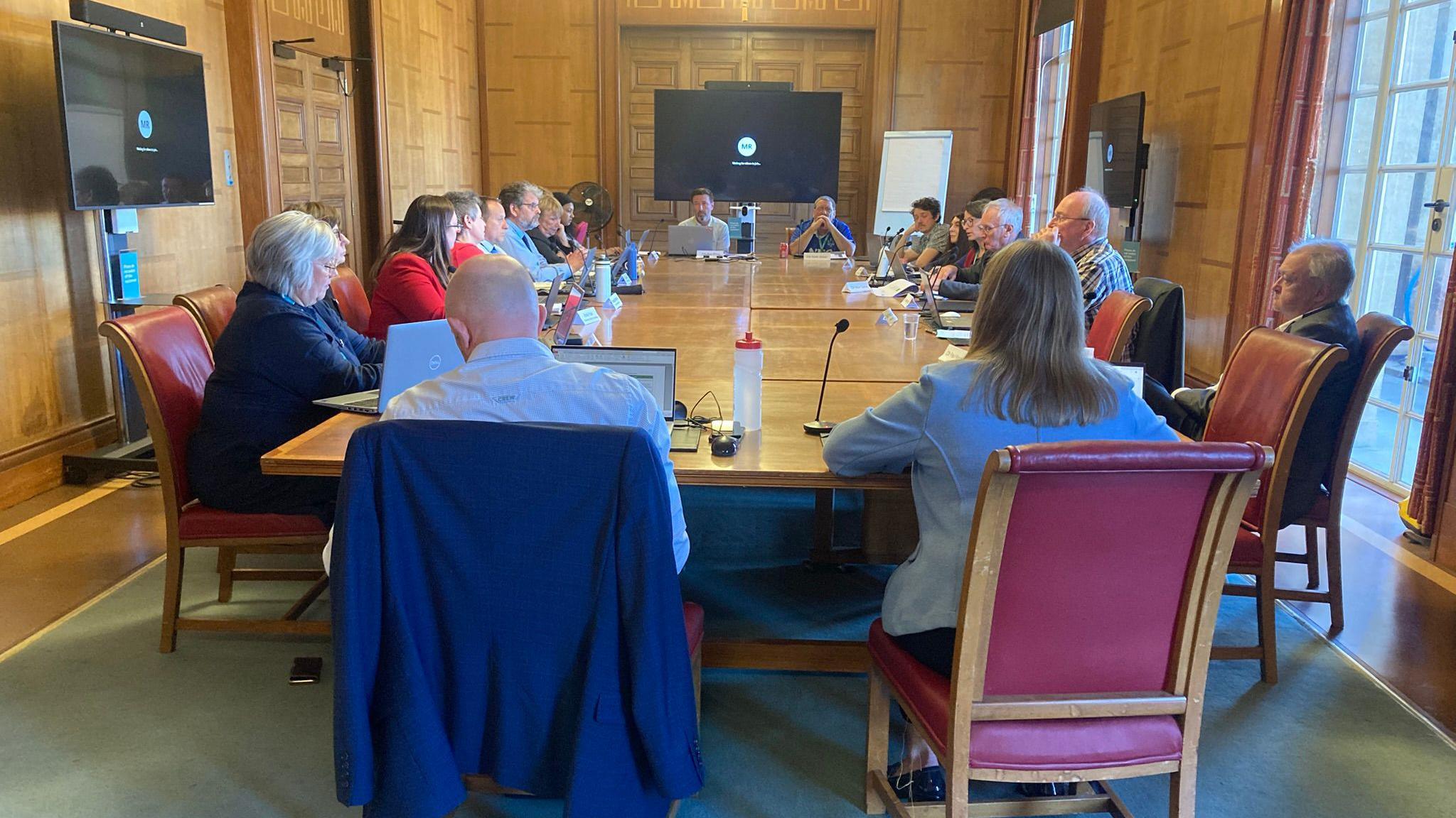 People sat around a large table in a committee room. The room has wood panels. There are three television screens positioned on the walls.