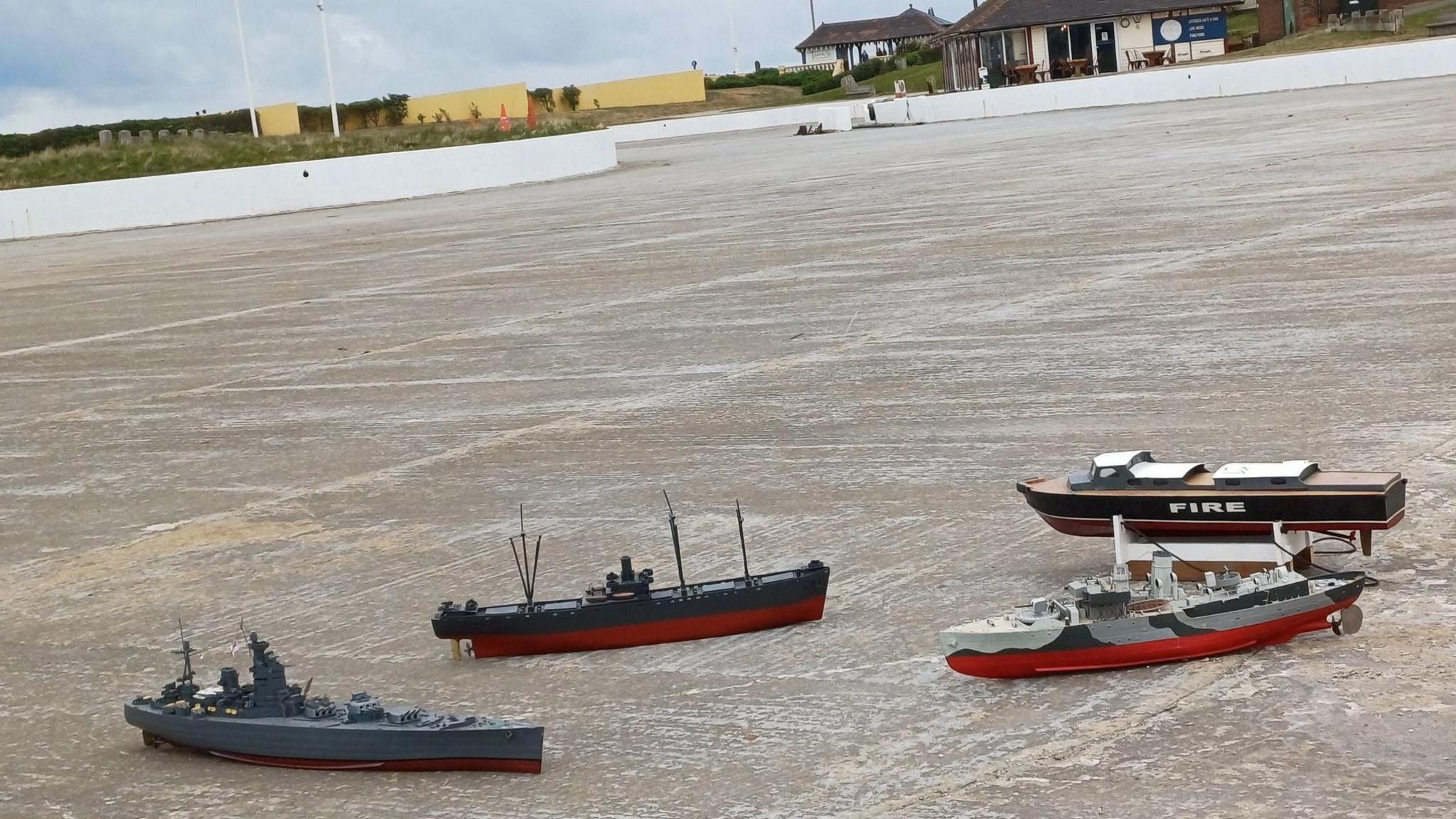 Four model boats laying on the concrete of the empty boating lake in Coatham, Redcar. The structure has a curving shape and is near the sea. The Boathouse and Terrace cafe is visible in the background.