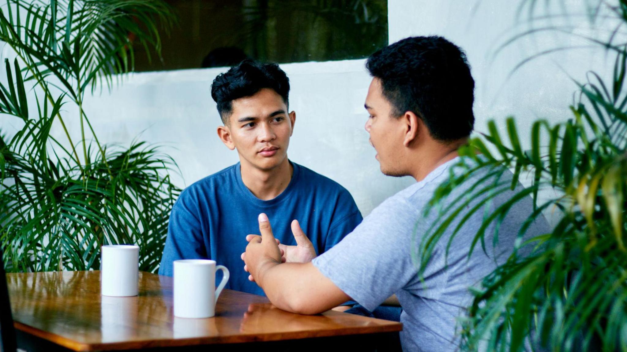 Two men with black hair sat at a brown table with two white mugs on top of it, green plants on either side of them. 