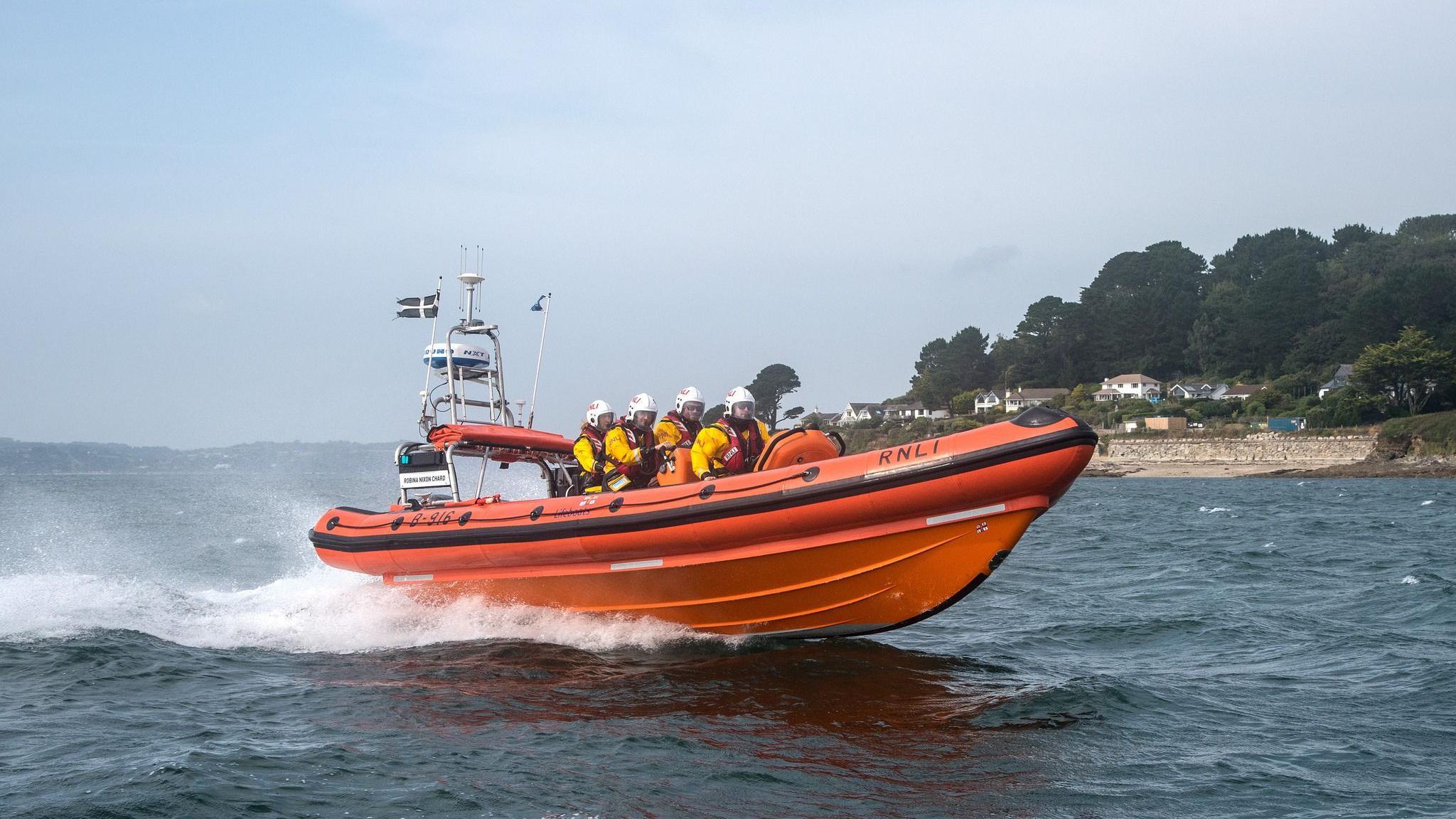 The front view of a orange lifeboat with four people on it. The lifeboat is moving on the water. The crew are wearing yellow life jackets. There is land to the right in the distance with large trees and houses.