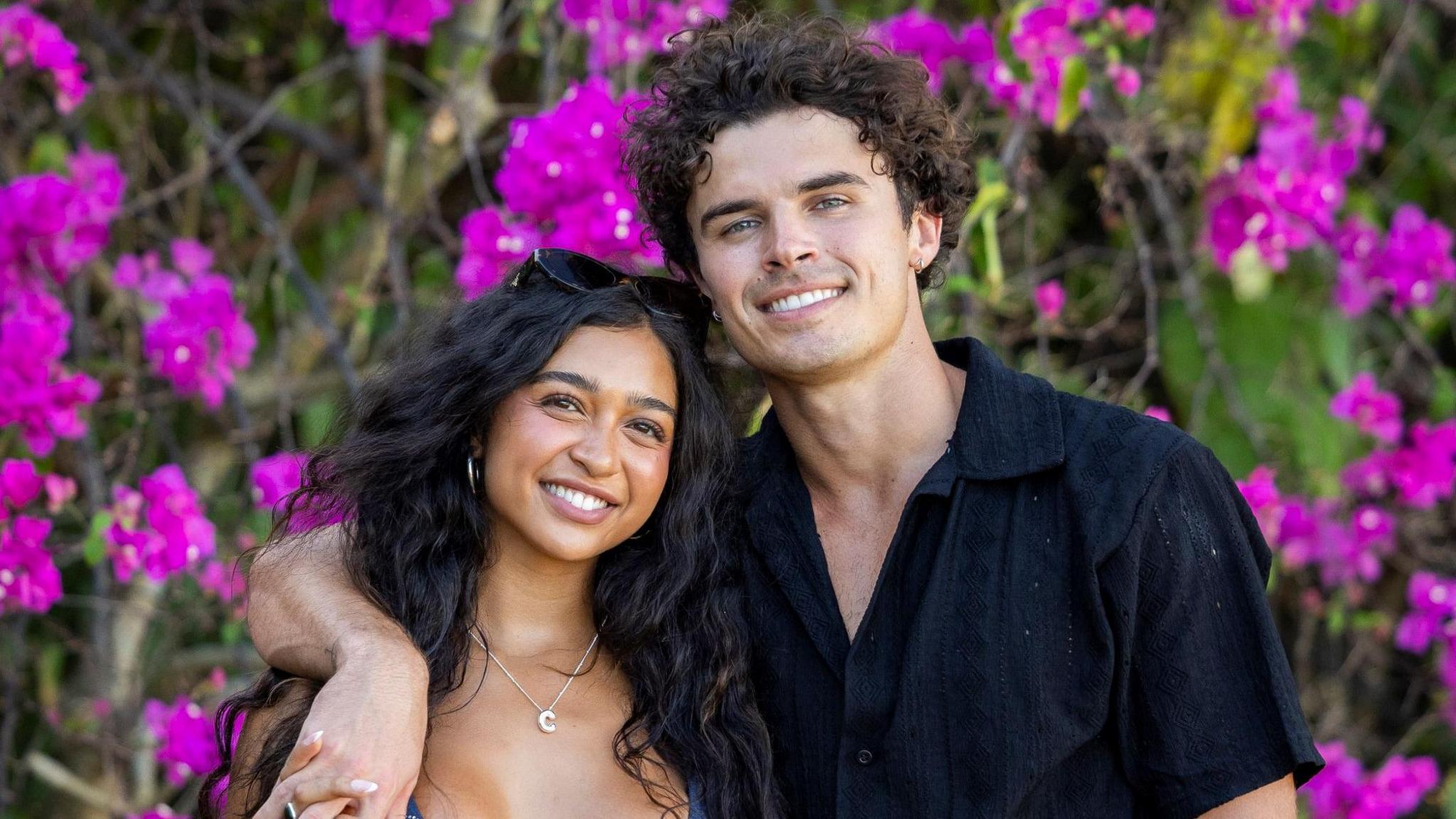 Cierra Ortega and Nic Vansteenberghe pictured in front of purple flowers in the Love Island USA villa. Cierra has long, curly, dark hair and smiles at the camera, her sunglasses perched in her hair. Nic has his arm around her shoulder, holding her hand. He has short curly hair and wears a black short-sleeved shirt.