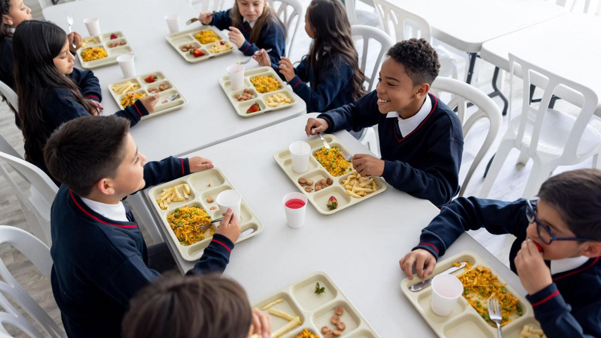 Children in school uniform eat school dinners from trays and are laughing and talking together 