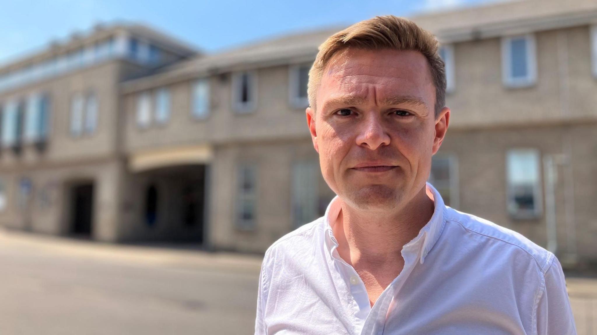 Will Quince wearing a white unbuttoned shirt on a sunny day. He has light brown hair and is looking at the camera with a slight frown.