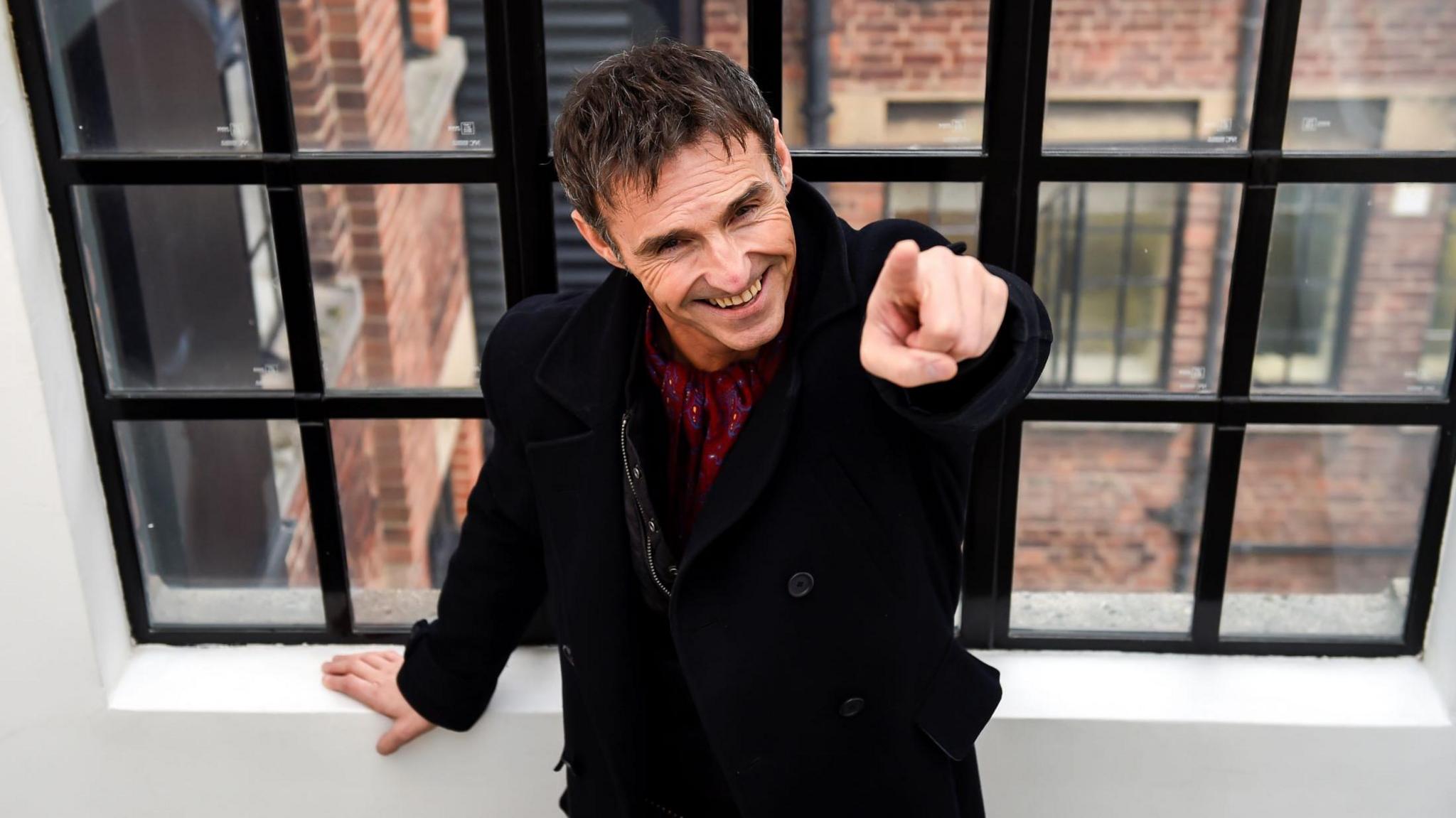 Marti Pellow points up to the camera with a cheeky smile while leaning on a windowsill. The image is taken from above and we can see an industrial-looking building outside. He is wearing a black wool coat and a burgundy scarf tucked in.