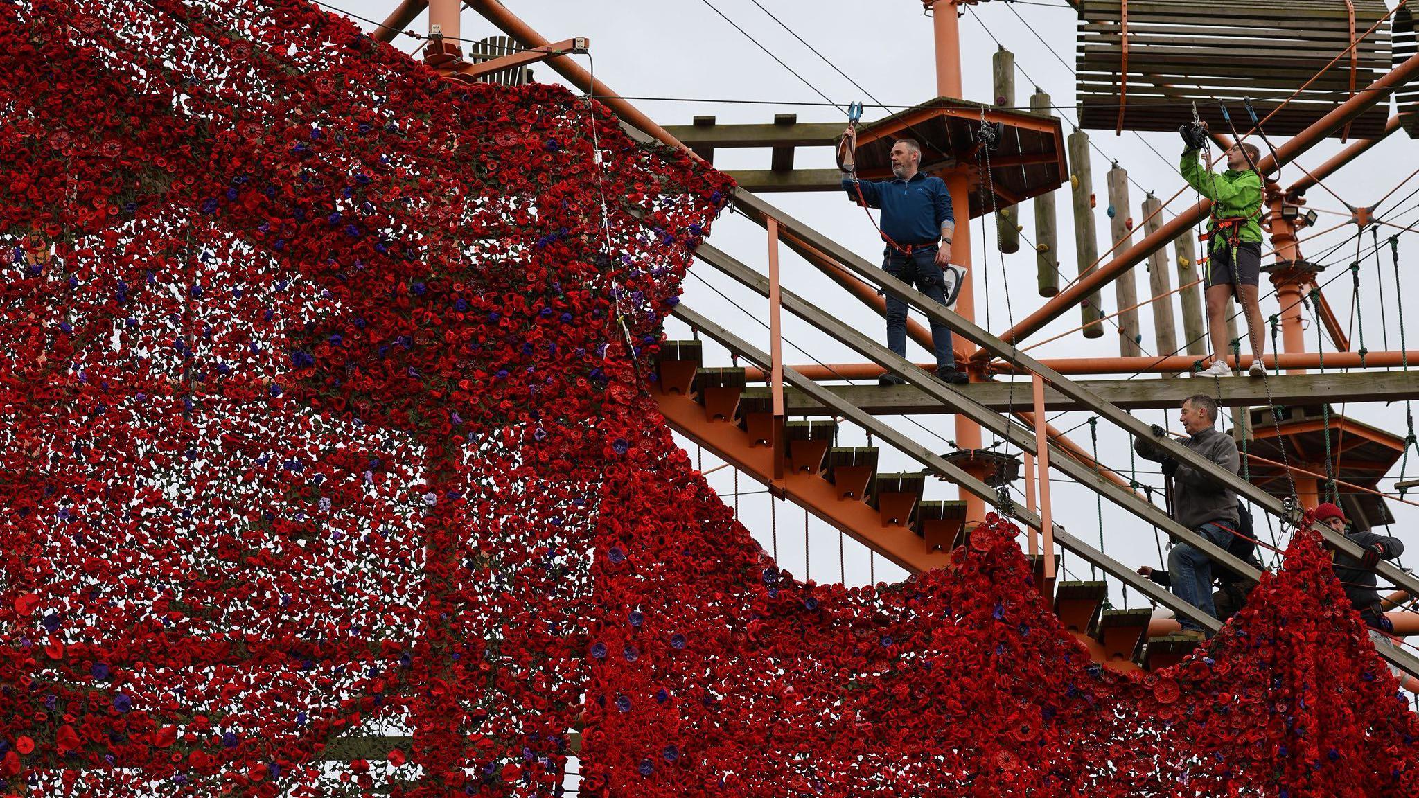 A sea of red with a few purple knitted poppies draped across a railing. One half of it is tied to the railing whilst four people can be seen working to secure the rest of the display on a high-wire attraction.