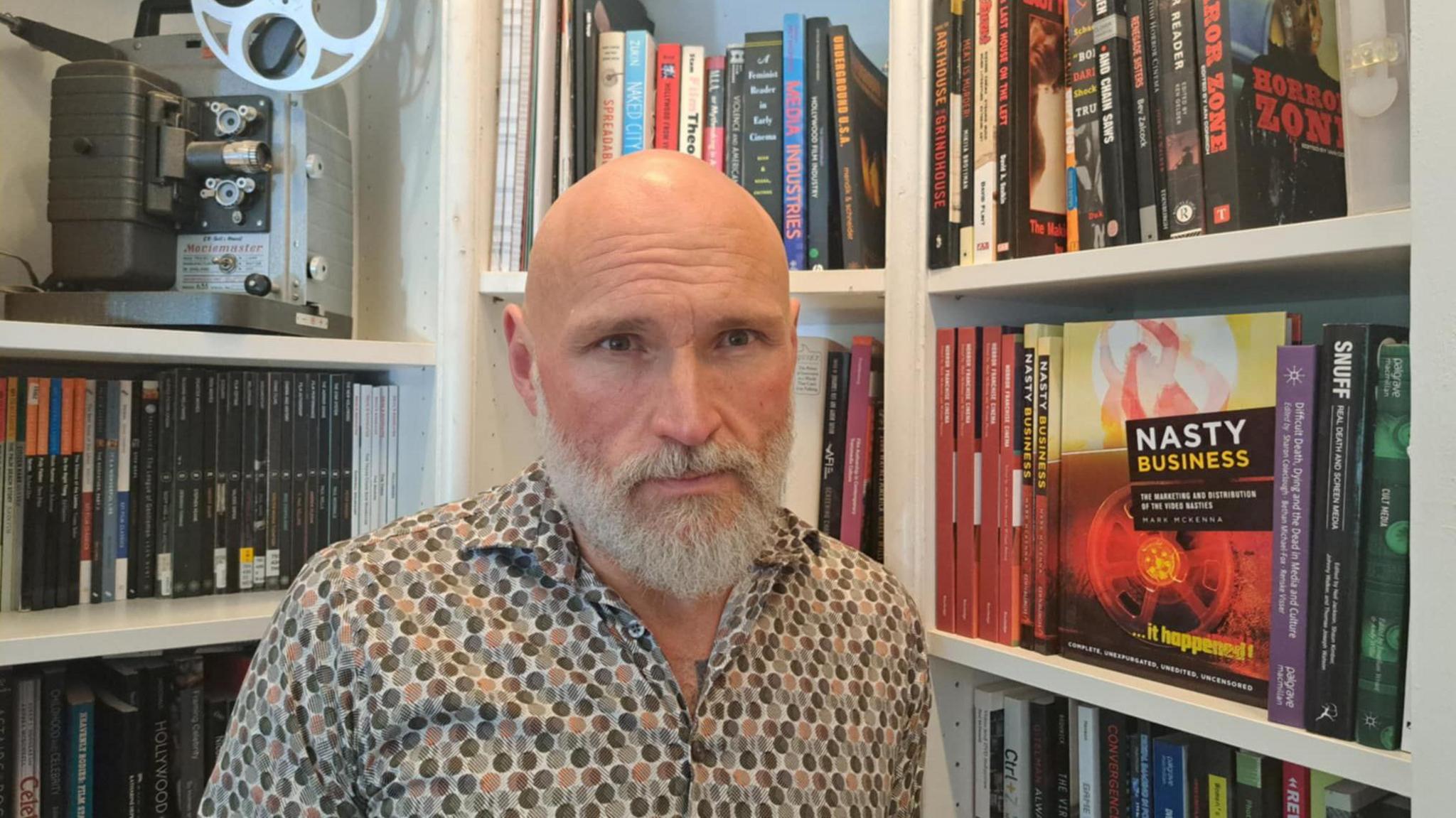 Image shows academic Dr Mark McKenna with bookshelves and books behind him and film equipment.