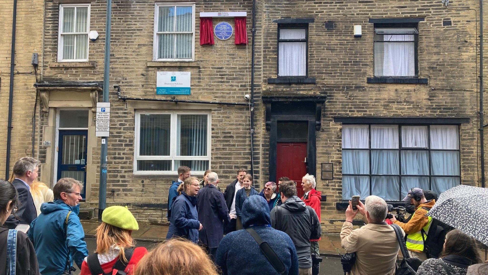 A crowd of people outside a terrace row of offices with a Blue Plaque on the outside wall on the first floor.