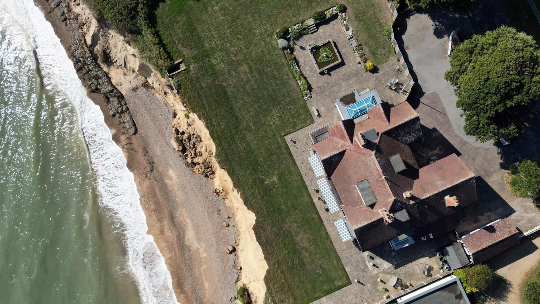 A photo taken aerially. It shows the sea on the left hand side, a narrow strip of beach, then bare cliff side and then a narrow strip of green grass. On the right hand side is a large house.