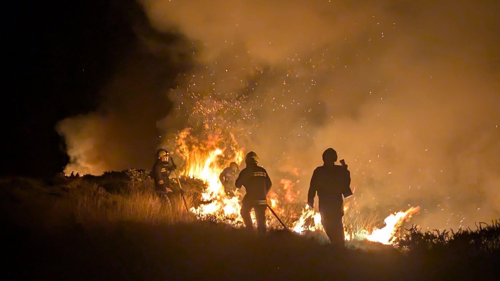 Three firefighters tackling a huge blaze on moorland. It is dark, and there are firefighters walking towards the orange flames. 