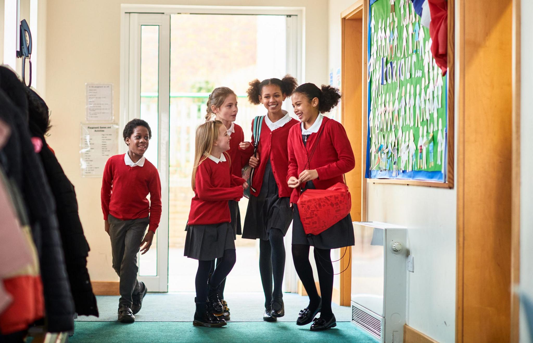 Five school children wearing red and black school uniform walk and laugh down a school corridoor.